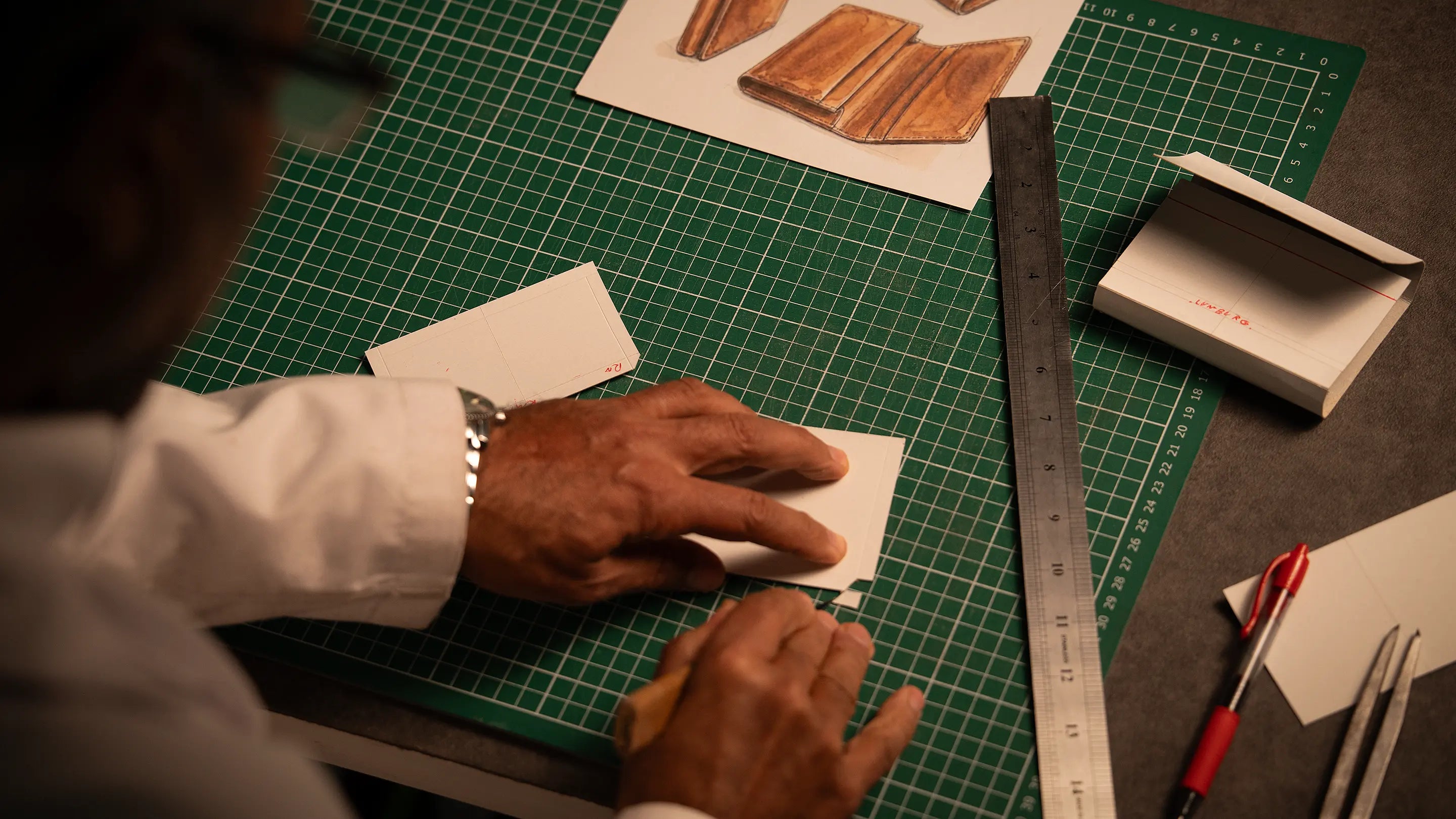 Artisan's hands at the Lunburg atelier working on a paper prototype alongside watercolor design renderings, steel ruler, and cutting mat, the origination phase of a Sovereign Commission where vision takes its first physical form.
