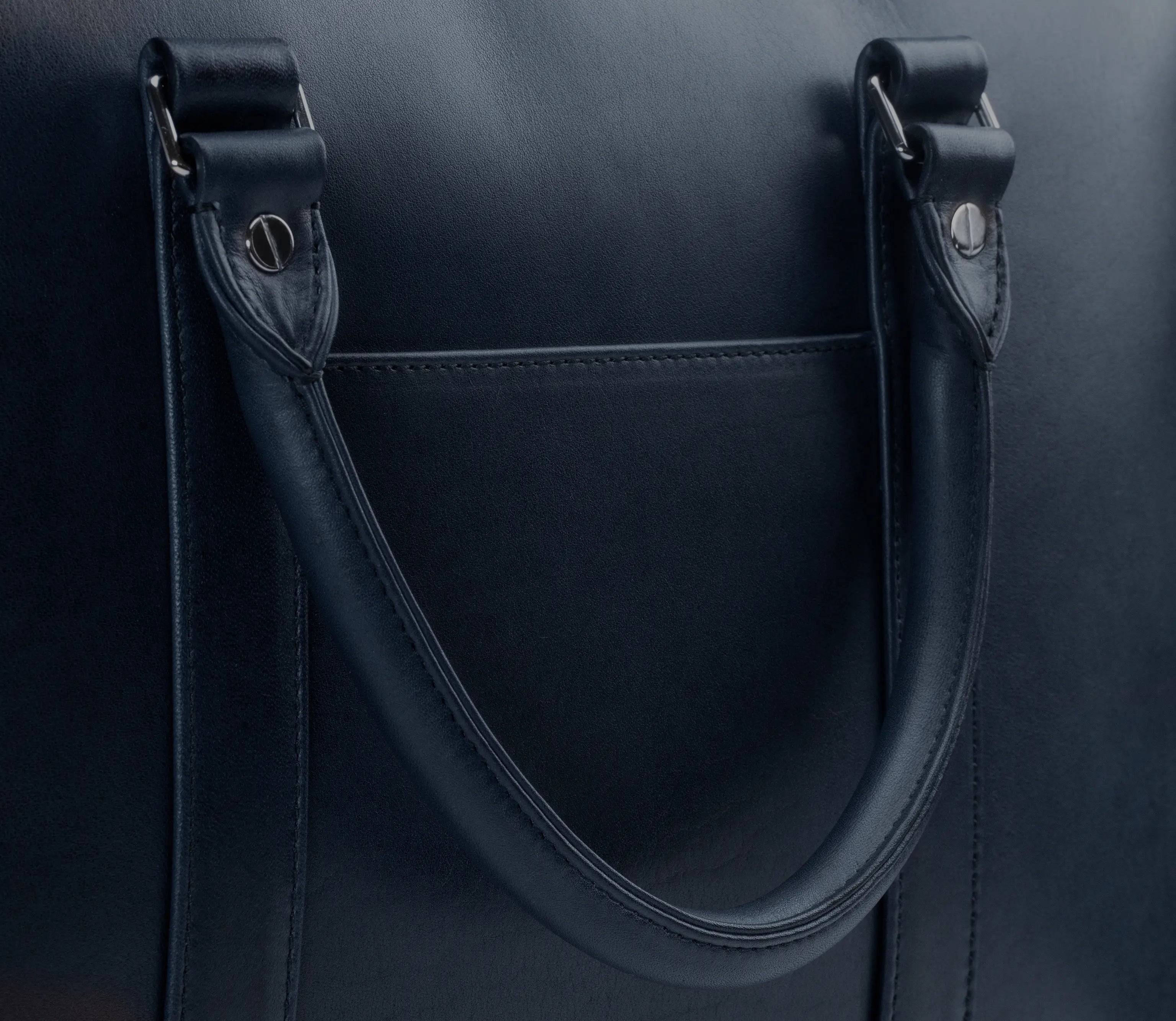 Detail of the Lunburg Opus Briefcase in Nocturnal Sapphire showing the folded leather handle loop, L-branded 316L stainless steel studs, slip pocket, and saddle stitching in vegetable-tanned Perpetual Leather (from Tempesti).