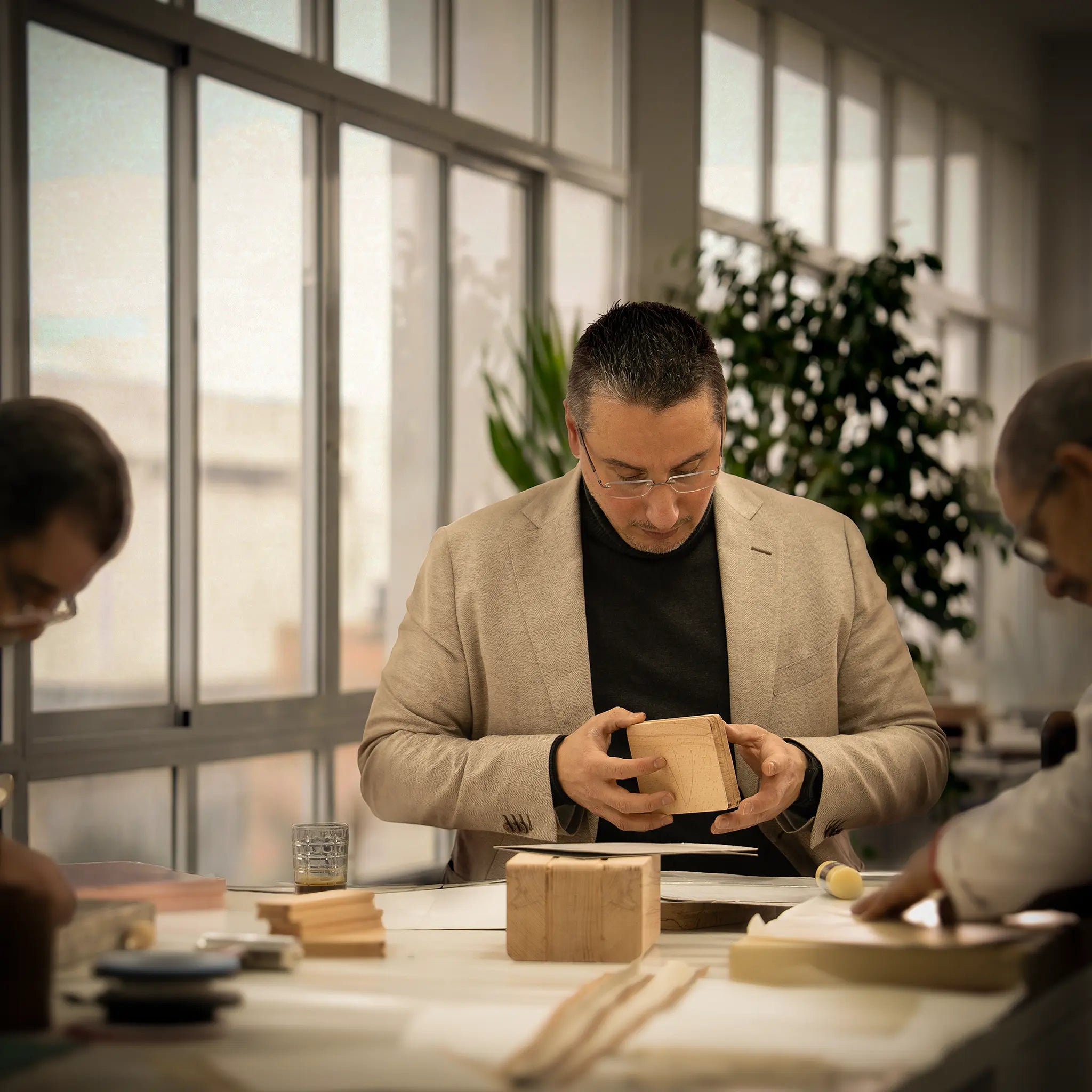 Lunburg founder Amien Marghich examining a leather prototype at the atelier workbench alongside master artisans, with wooden lasts, pattern templates, and traditional tools visible, natural window light illuminating the workspace in the Fes atelier.