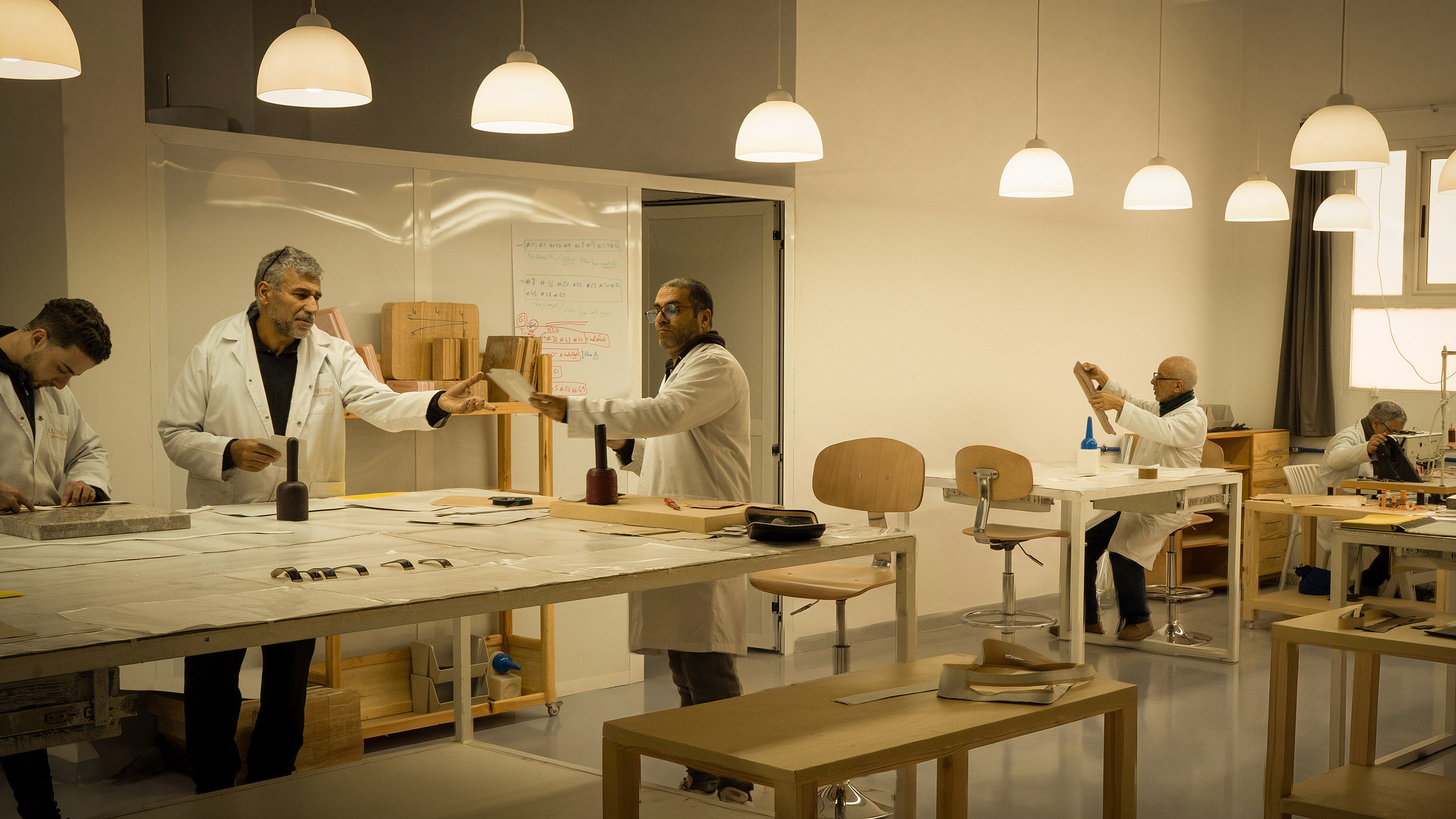 Wide view of the Lunburg atelier in Fes, with master artisans in white coats working at individual stations surrounded by wooden lasts, marble slabs, rempliage hammers, and traditional leatherworking tools, preserving twelve centuries of Fassi haute maroquinerie.