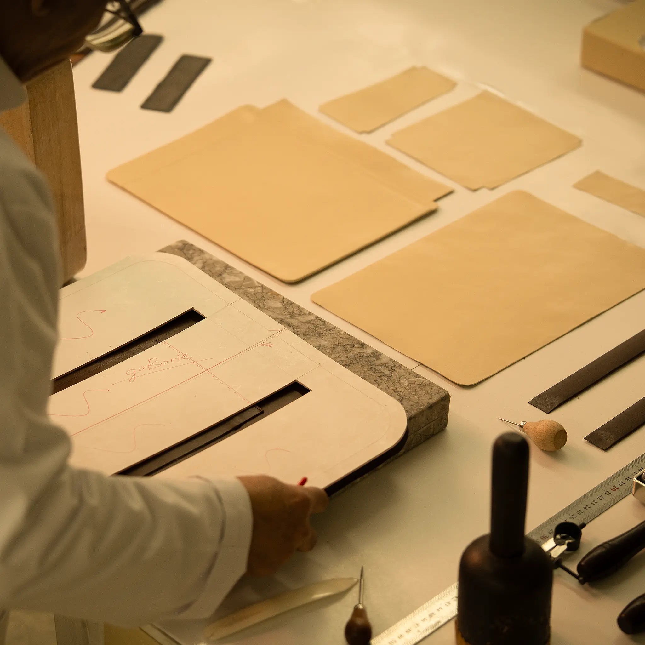 Artisan's hand at the Lunburg atelier reviewing pattern templates alongside cut leather panels, marble slab, rempliage hammer, and precision tools, the beginning of a bespoke commission where patron specifications meet craft execution.