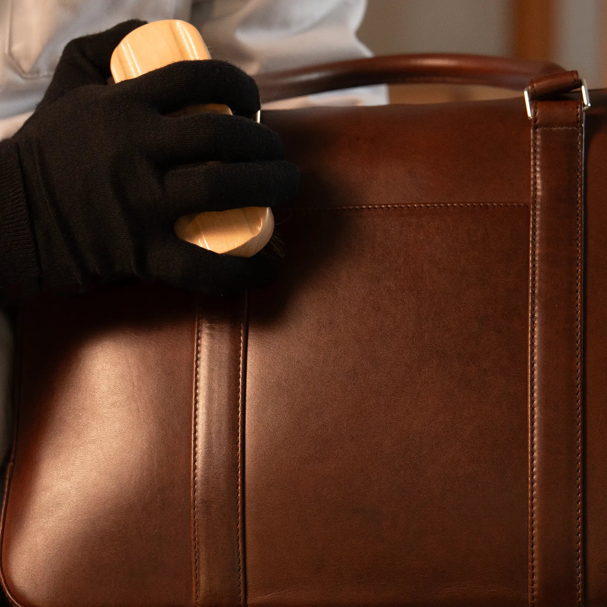 Gloved hand brushing the surface of a Lunburg Opus Briefcase in Deep Mahogany with a natural bristle brush at the Fes atelier, reviving the natural resilience of vegetable-tanned Perpetual Leather with folded edge construction.