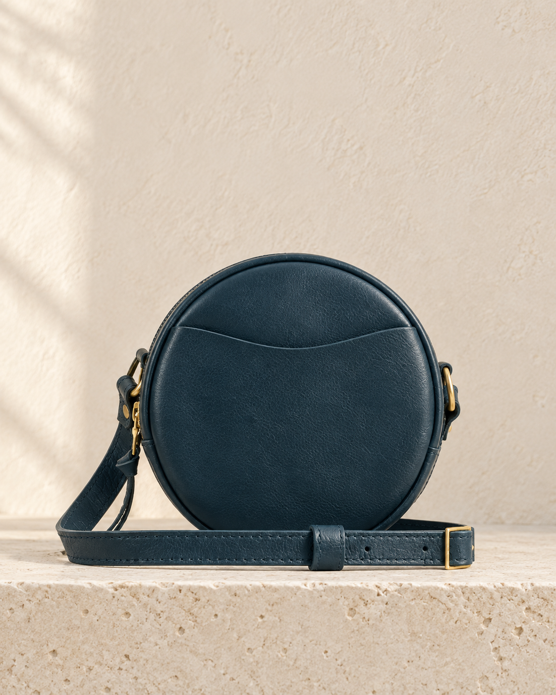 Illustrative circular crossbody with raised perimeter piping, a curved zip opening, front slip pocket and ring-mounted strap.
