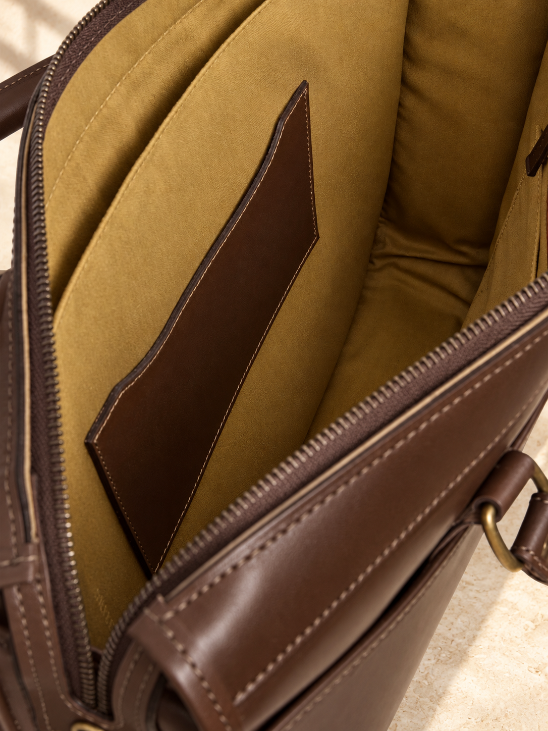 Partial interior of the illustrative briefcase showing ochre lining, a broad sleeve or divider, a large sewn leather patch and the zip rim.