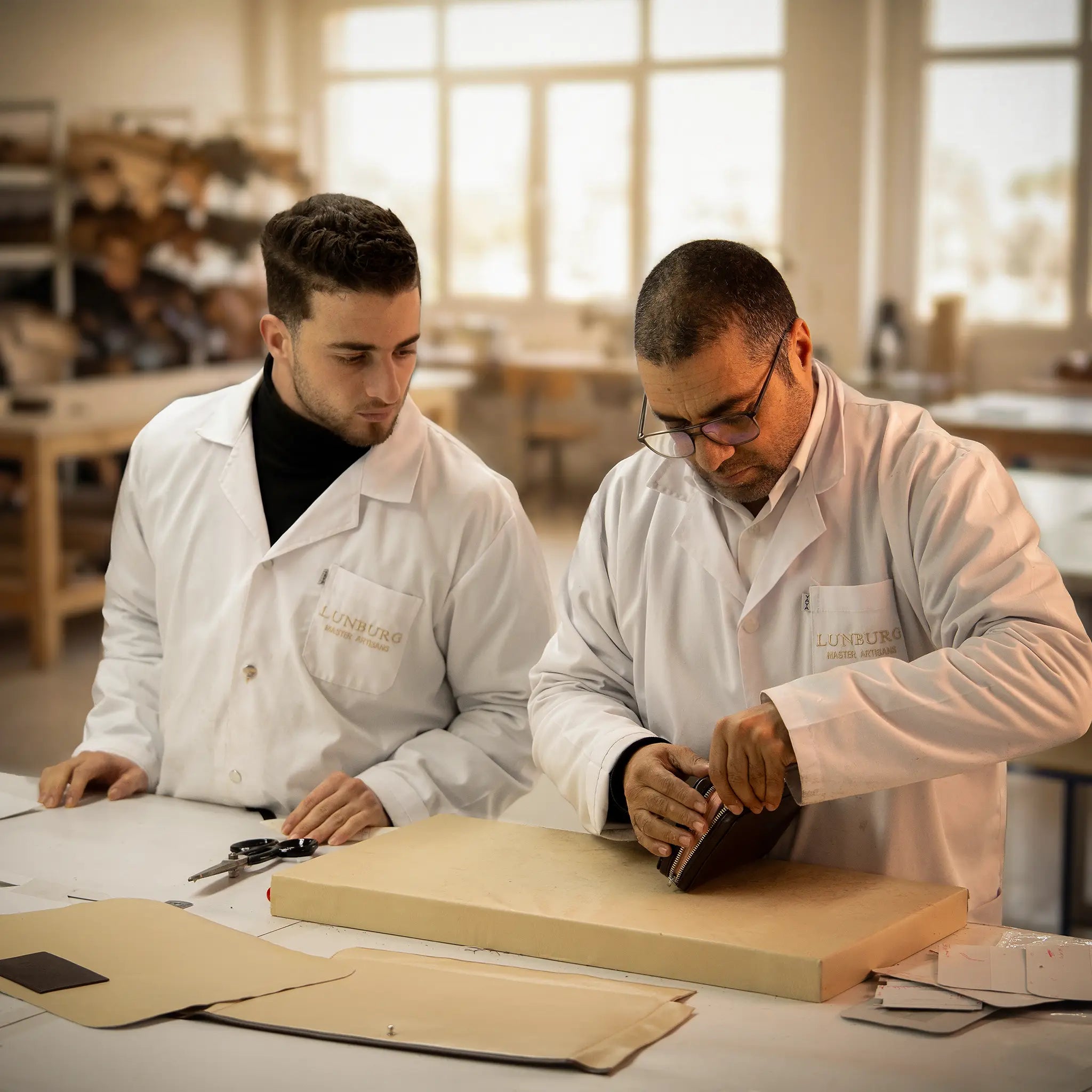 A Lunburg Master Artisan mentoring a young apprentice at the Fes atelier, both in Lunburg white coats, working together on a leather piece with pattern templates and tools on the workbench, the transmission of twelve centuries of Fassi heritage knowledge.