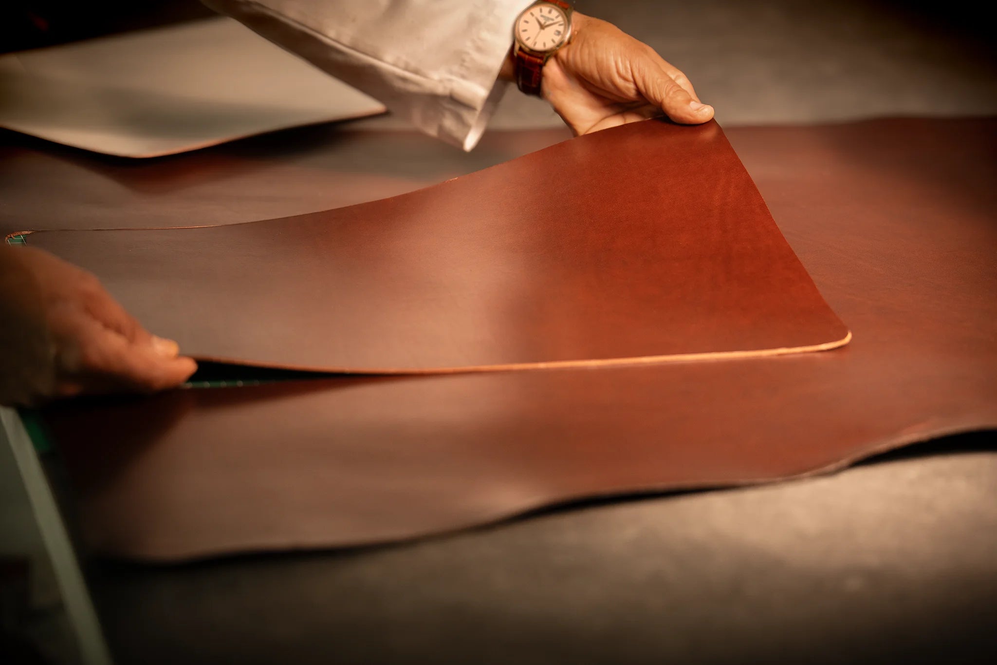 A Master Artisan lifts a freshly cut panel of vegetable-tanned Perpetual Leather (from Tempesti) from a full hide spread across the cutting table at the Fes atelier, the single-cut selection moment that determines the unique grain character of each Memoir portfolio.