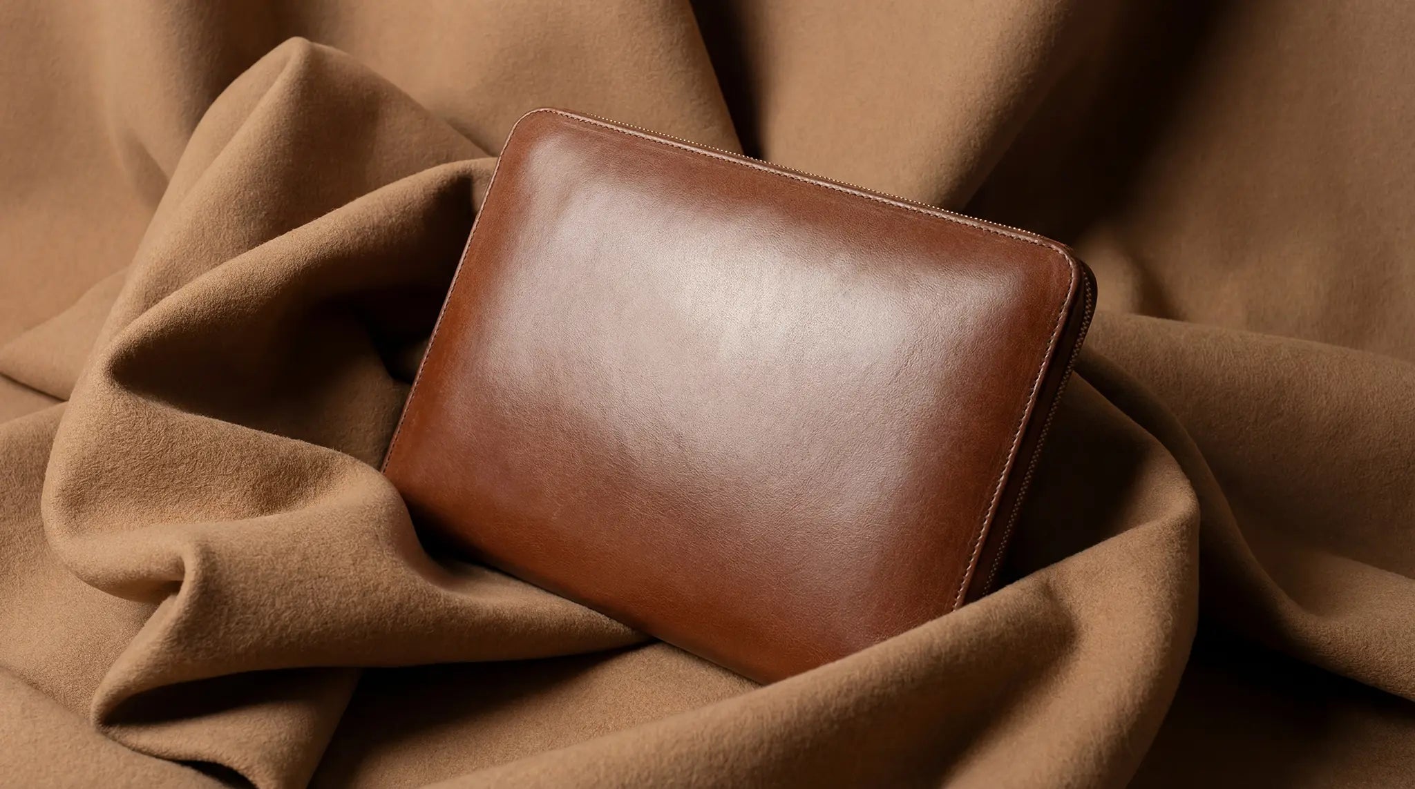 Lunburg Memoir portfolio in Heritage Amber vegetable-tanned Perpetual Leather (from Tempesti) rests closed on draped camel fabric, soft directional light revealing the natural grain across the single-cut exterior panel with saddle stitching along the edges — the singular object embodying Visual Silence.