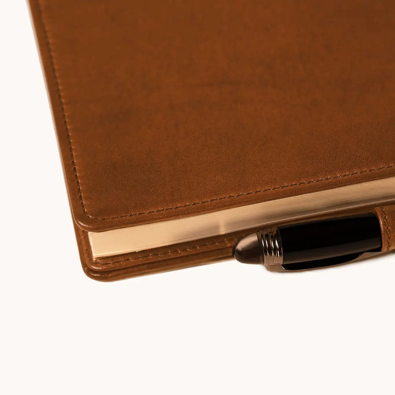 Lunburg Memoir Notebook Cover with Pen Loop in Heritage Amber with an A5 notebook inserted and a pen held in the leather loop, resting on a table at a three-quarter angle showing the cover's profile, spine, and page block, in vegetable-tanned Perpetual Leather (from Tempesti) with folded edge construction.