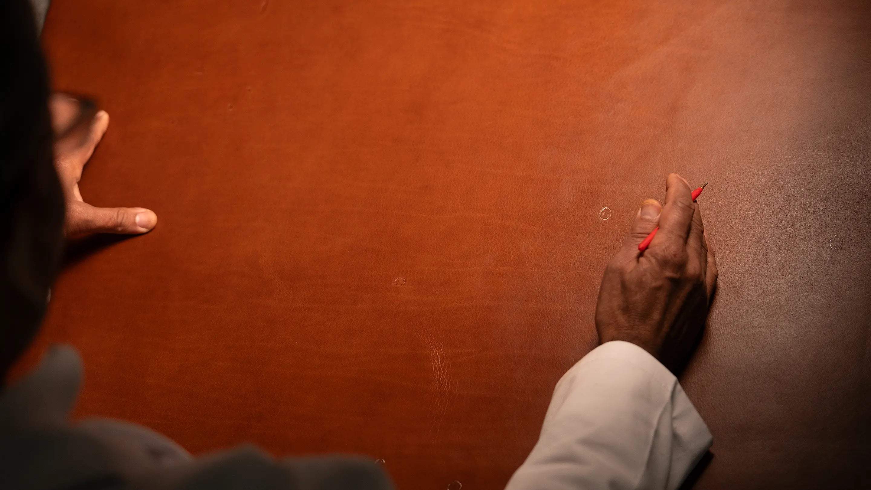 Master Artisan's hand marking a full hide of vegetable-tanned Perpetual Leather with a red marking tool, selecting from the double-bend core where each grain is as singular as a fingerprint, no two pieces ever alike.