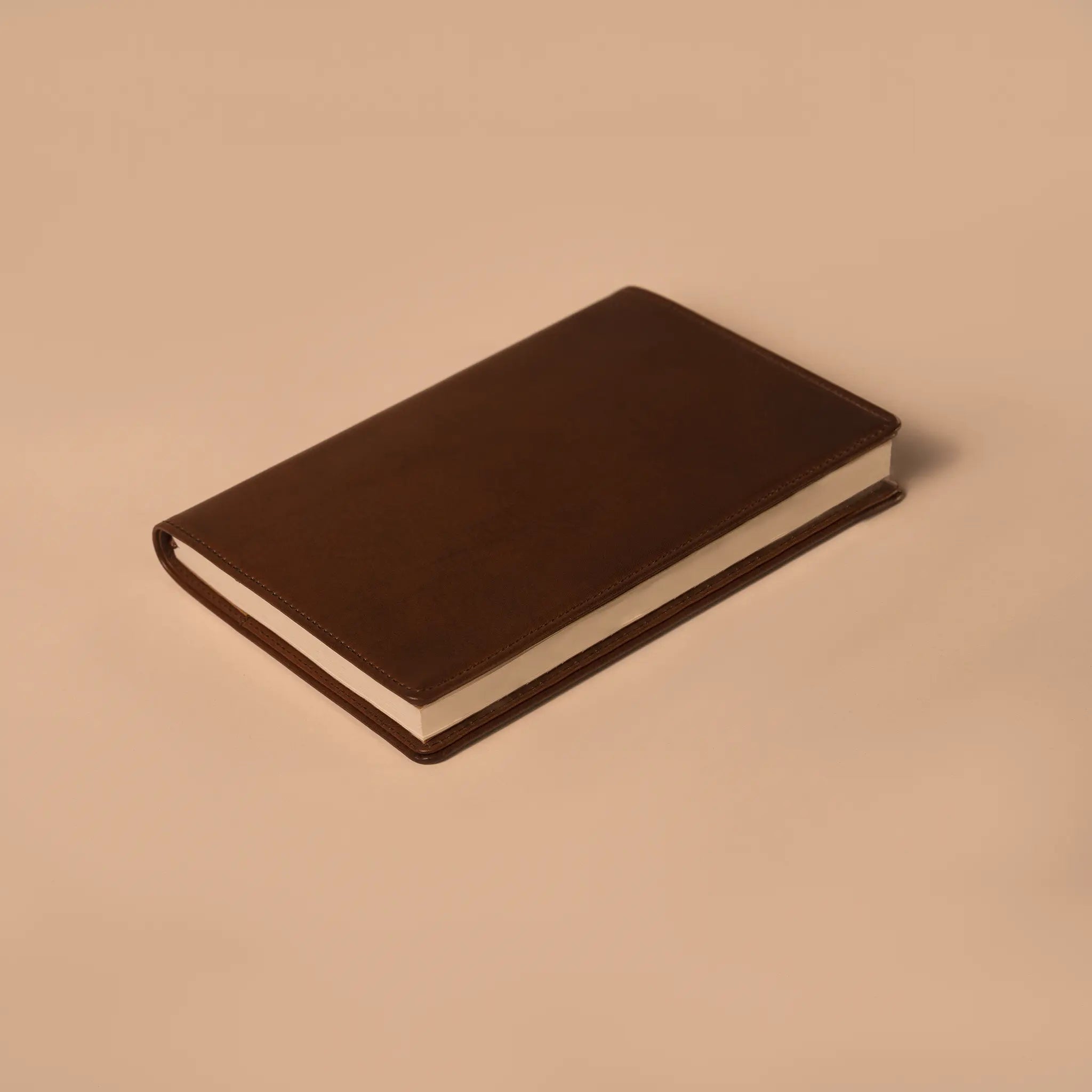 Lunburg Memoir Pocket Notebook Cover in Deep Mahogany with a pocket notebook inserted, resting on a table at a three-quarter angle showing the leather cover's profile, spine, and page block, in vegetable-tanned Perpetual Leather (from Tempesti) with folded edge construction.