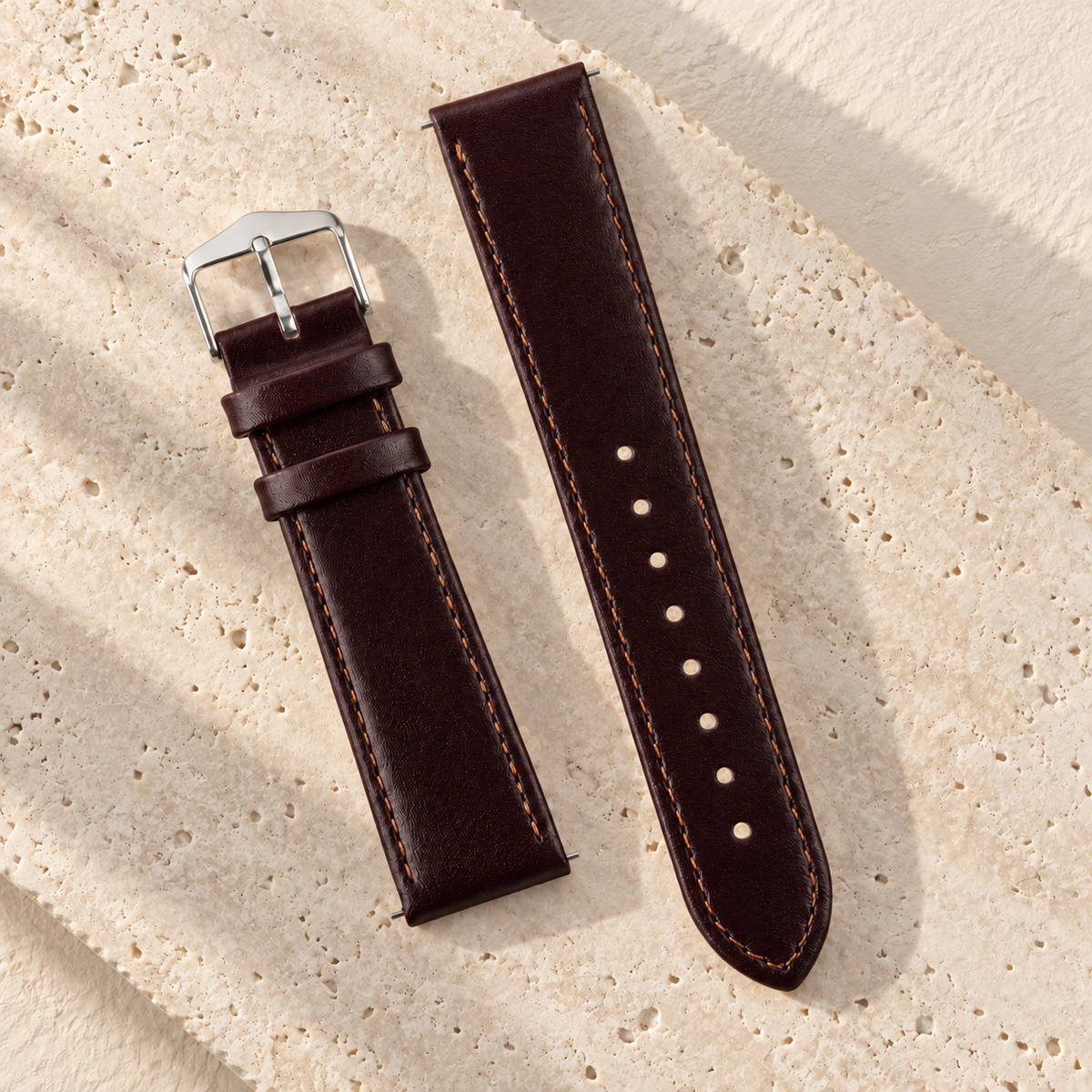 Illustrative tapered tang-buckle watch strap, view 1: two burgundy strap pieces with tapered perforated end, tang buckle, two keepers and perimeter stitching.