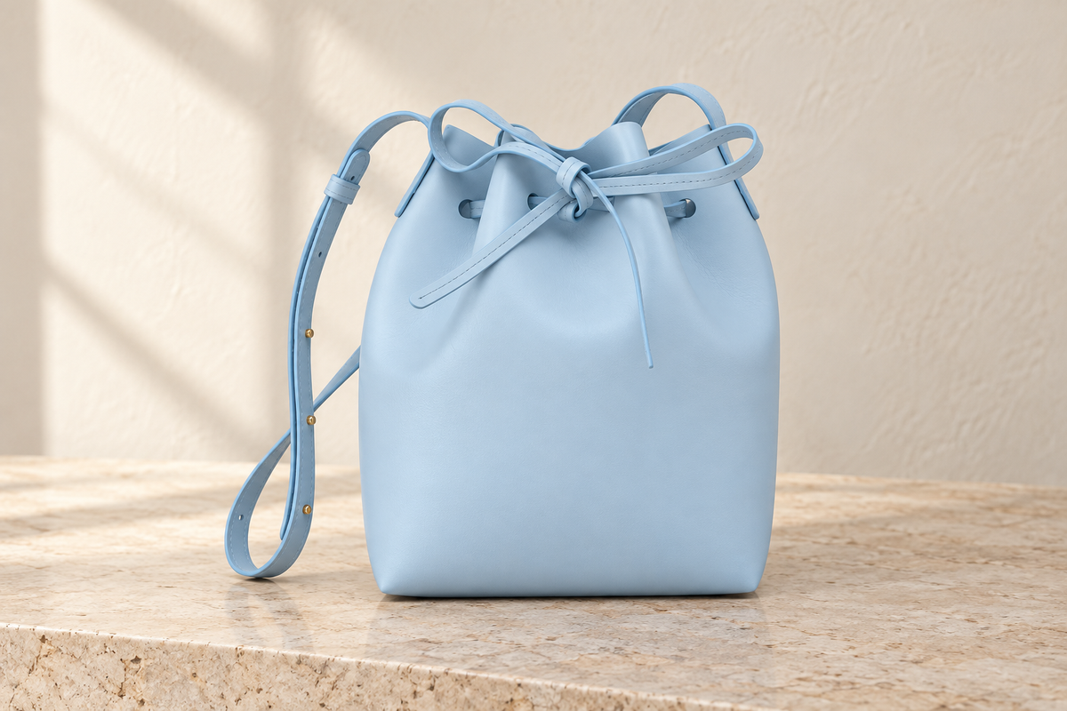 Illustrative powder-blue bucket bag with a gathered neck, tied flat drawstring and narrow shoulder strap with side studs.
