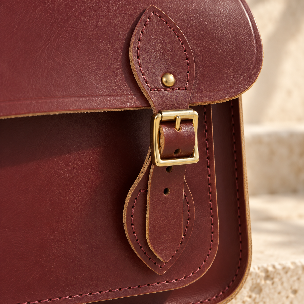Detail of the illustrative satchel showing a buckle strap, leaf-shaped stitched and riveted mount, and outlined stitch rows.