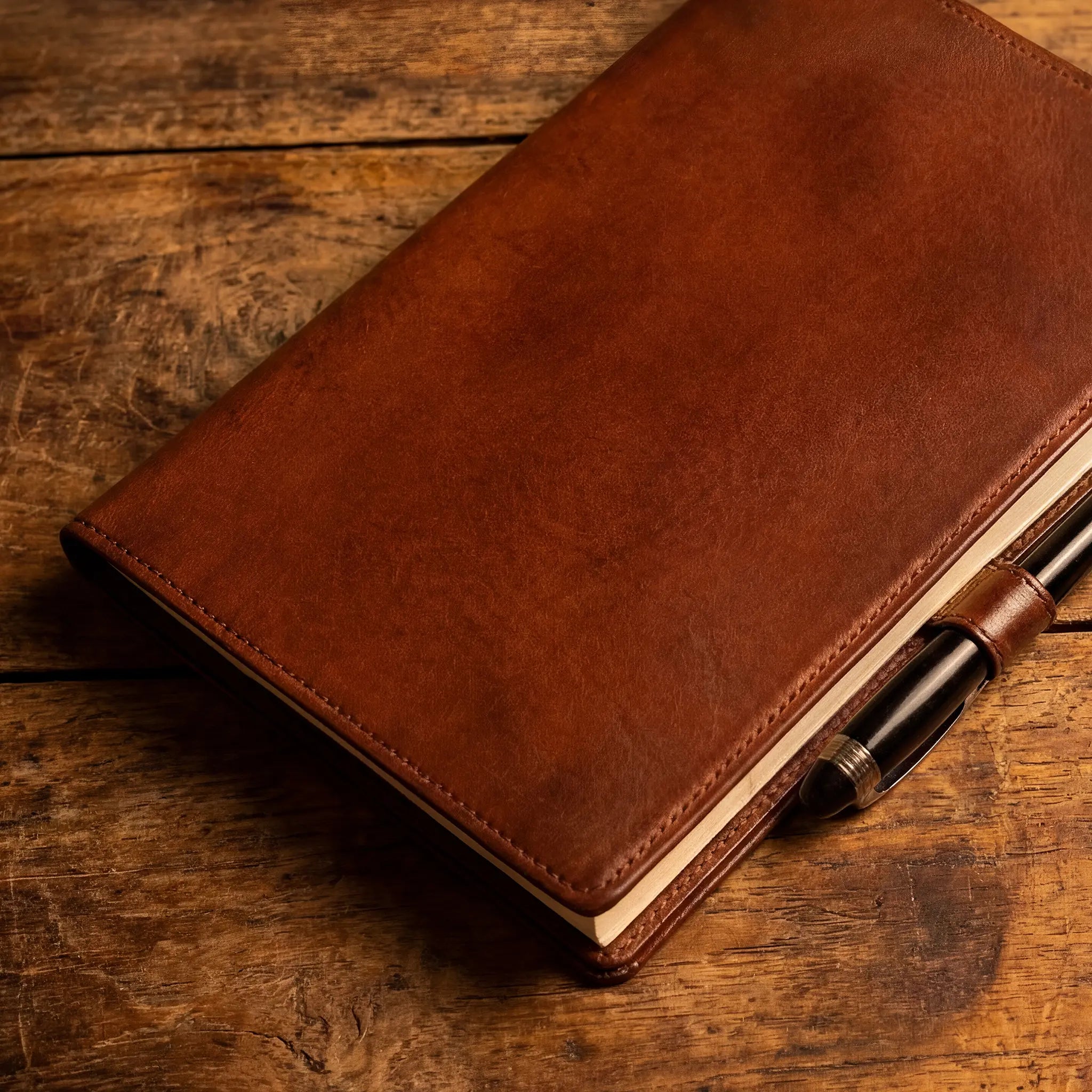 Lunburg Memoir Notebook Cover in Heritage Amber resting on aged wood with a fountain pen in the leather pen loop, showing the natural patina and softening of vegetable-tanned Perpetual Leather with folded edge construction, the signs of life the Fifty-Year Warranty celebrates.