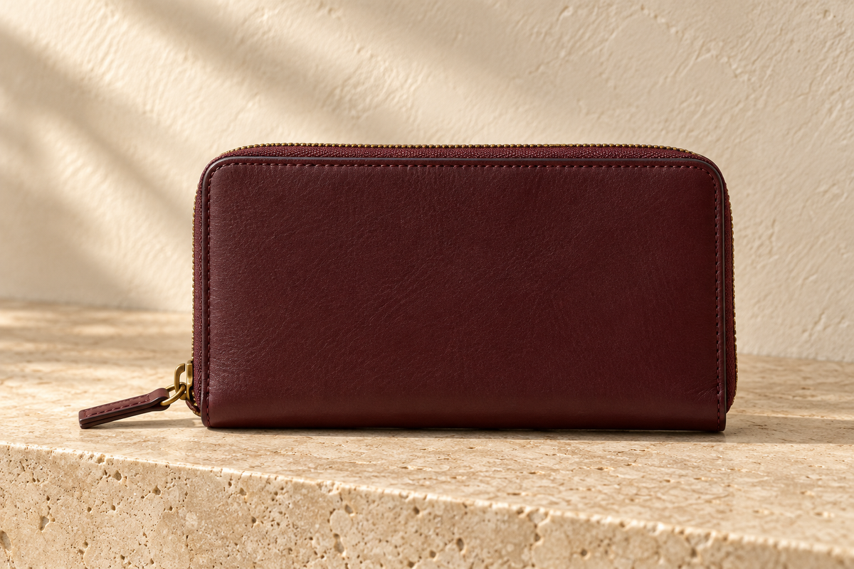 Illustrative zip-around divided wallet — long rectangular zip wallet with rounded upper corners, plain burgundy face, visible perimeter stitch line and projecting stitched zip pull.