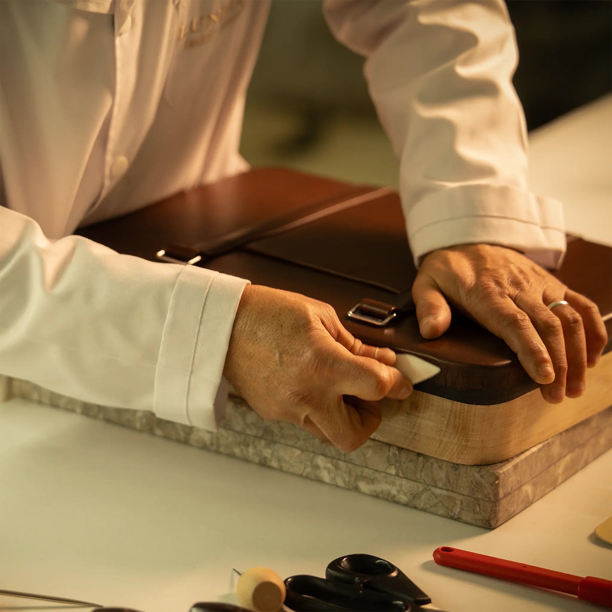 Master artisan's hands at the Lunburg atelier assembling a leather piece on the forme en bois wooden last over a marble slab, with traditional tools nearby, executing the folded edge construction of Rempliage in vegetable-tanned Perpetual Leather.
