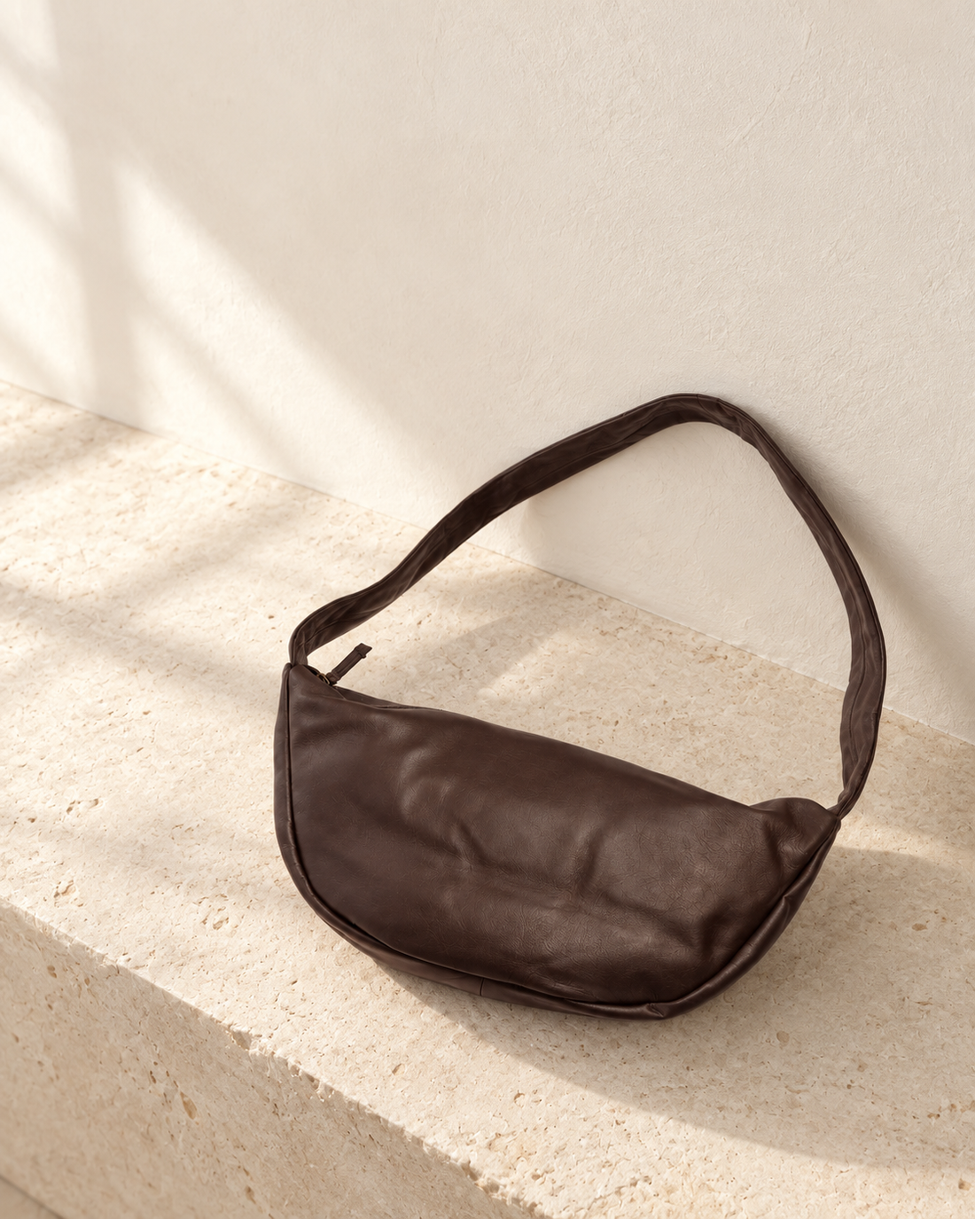 Illustrative crescent shoulder bag with a soft curved body, broad lower gusset and wide strap attached directly at the ends.
