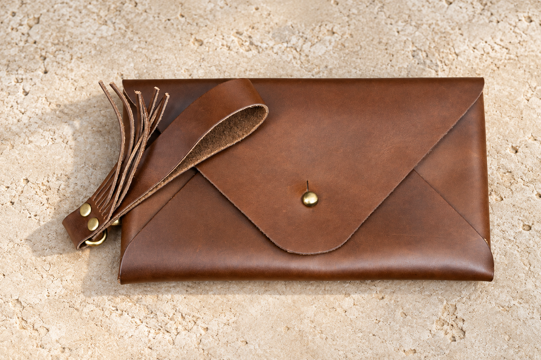 Illustrative envelope wristlet with a pointed-round flap, button-stud closure and clipped wrist loop with fringe.
