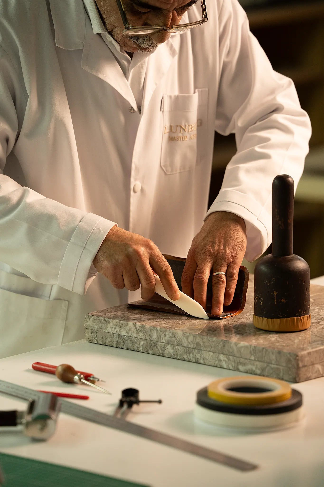 Lunburg Master Artisan folding a leather edge with a bone folder on a marble slab at the Fes atelier, rempliage hammer alongside, executing the Rempliage technique with the surgical precision inherited from Andalusian master leatherworkers who brought nine centuries of refinement to Fes after the fall of Granada in 1492.