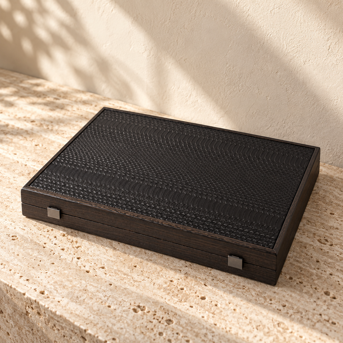 Illustrative folding backgammon case, view 1: closed rectangular folding case with dark scale-pattern surface, perimeter frame and two metal-coloured front clasps.