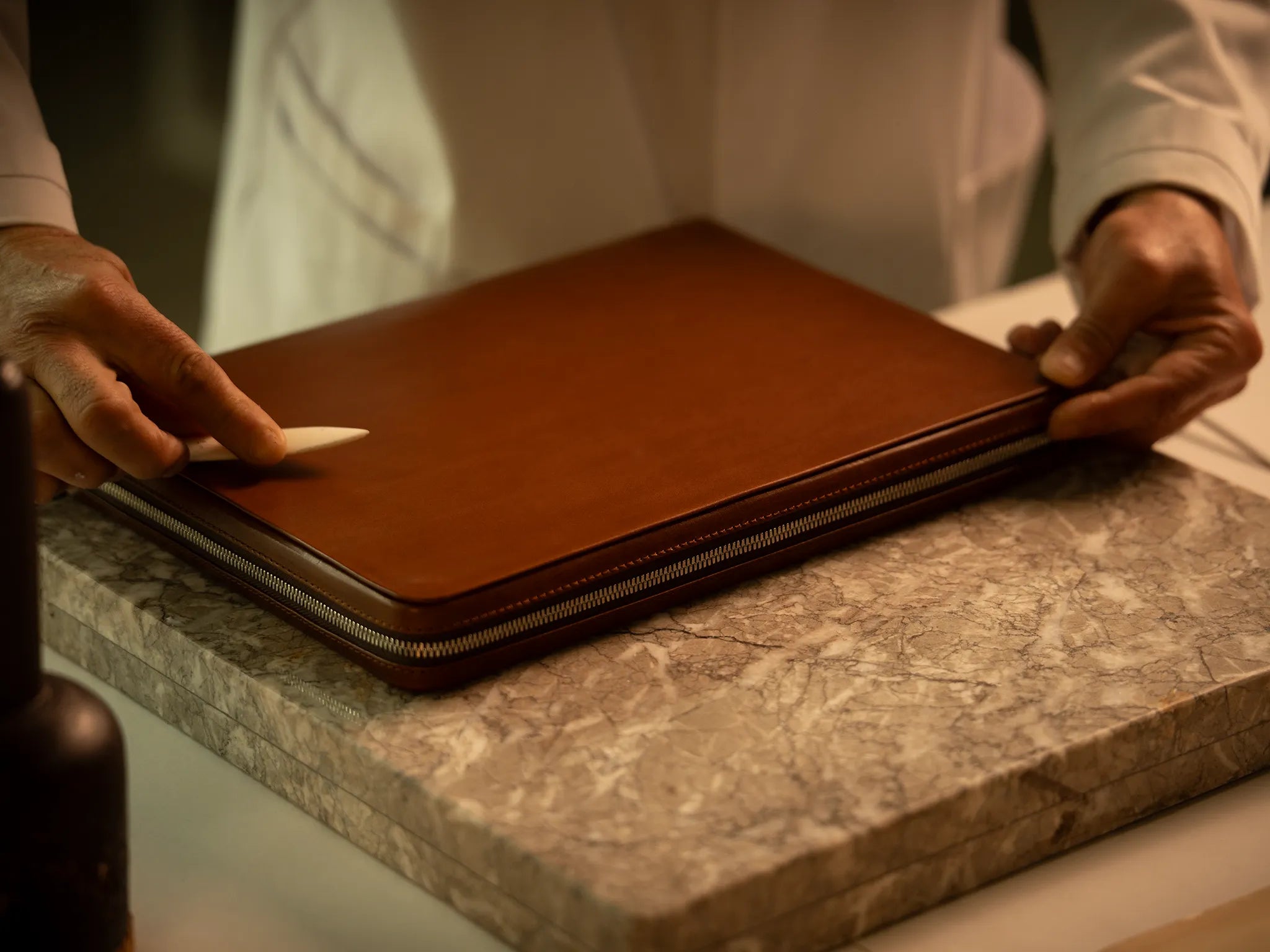 A Master Artisan uses a bone folder to press the vegetable-tanned Perpetual Leather (from Tempesti) surface of a Memoir portfolio in Heritage Amber at the Fes atelier, the YKK Excella zipper visible along the perimeter as the piece rests on a marble slab beside traditional hand tools.