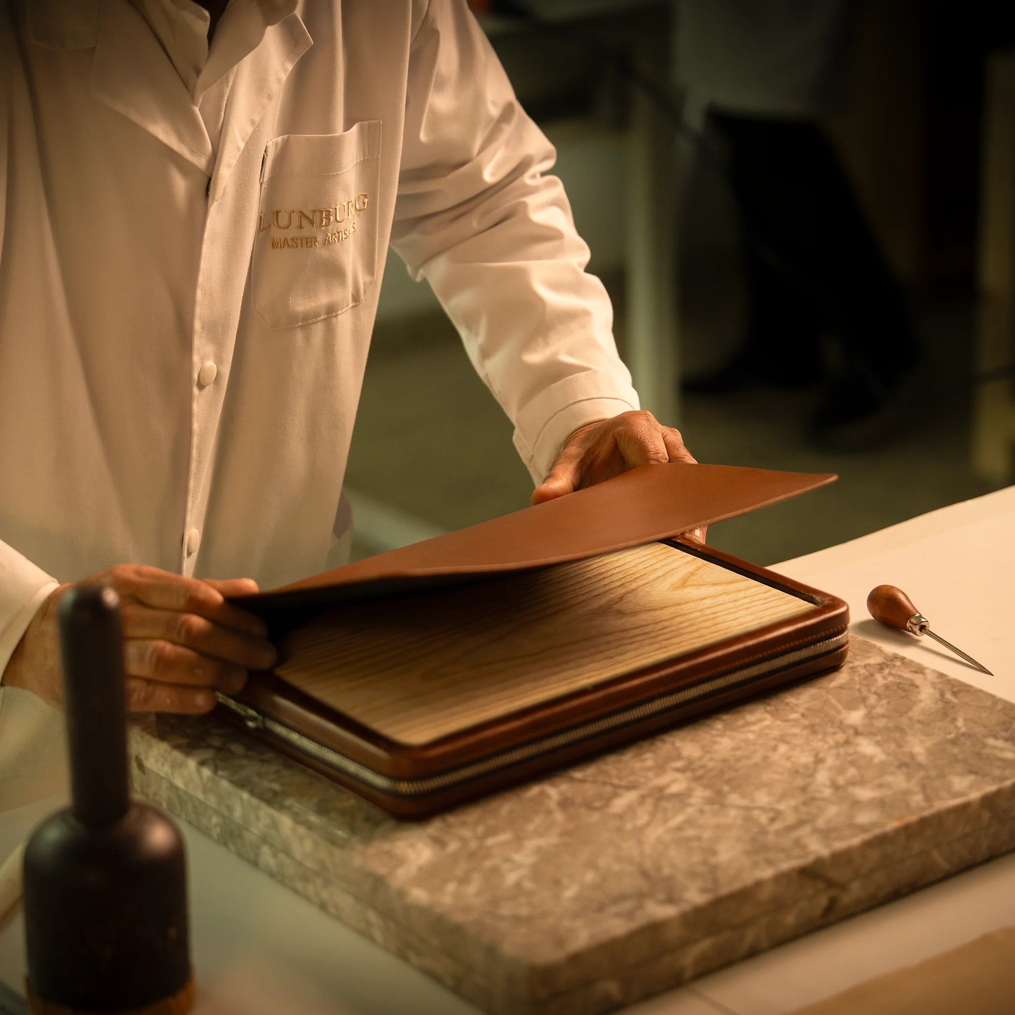 A Lunburg Master Artisan layers vegetable-tanned Perpetual Leather (from Tempesti) over a forme en bois at the Fes atelier, the wooden form imposing dimensional memory upon the Memoir's construction, with an awl and maul resting on the marble slab alongside the Heritage Amber assembly.