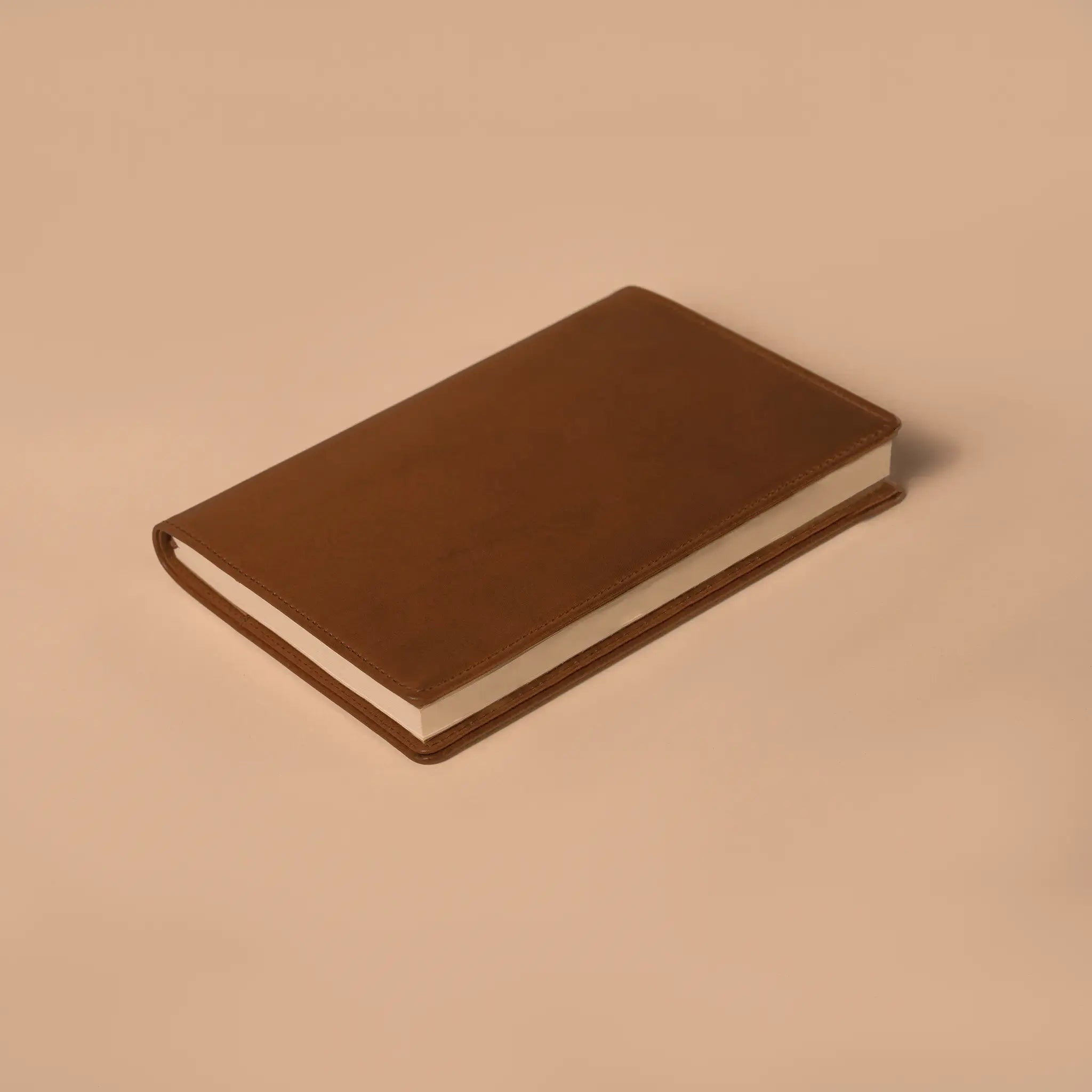 Lunburg Memoir Notebook Cover in Heritage Amber with an A5 notebook inserted, resting on a table at a three-quarter angle showing the leather cover's profile, spine, and page block, in vegetable-tanned Perpetual Leather (from Tempesti) with folded edge construction.