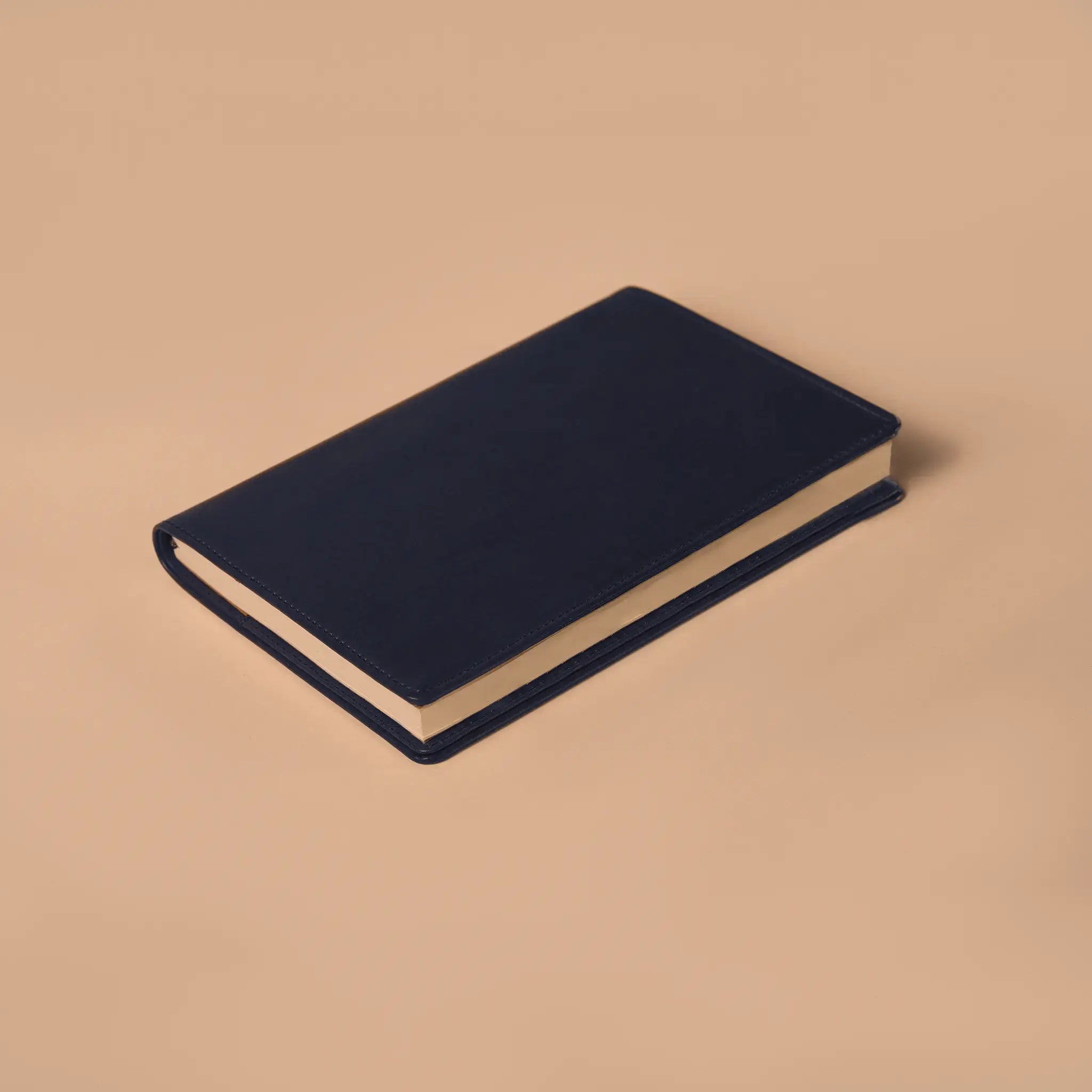 Lunburg Memoir Notebook Cover in Nocturnal Sapphire with an A5 notebook inserted, resting on a table at a three-quarter angle showing the leather cover's profile, spine, and page block, in vegetable-tanned Perpetual Leather (from Tempesti) with folded edge construction.