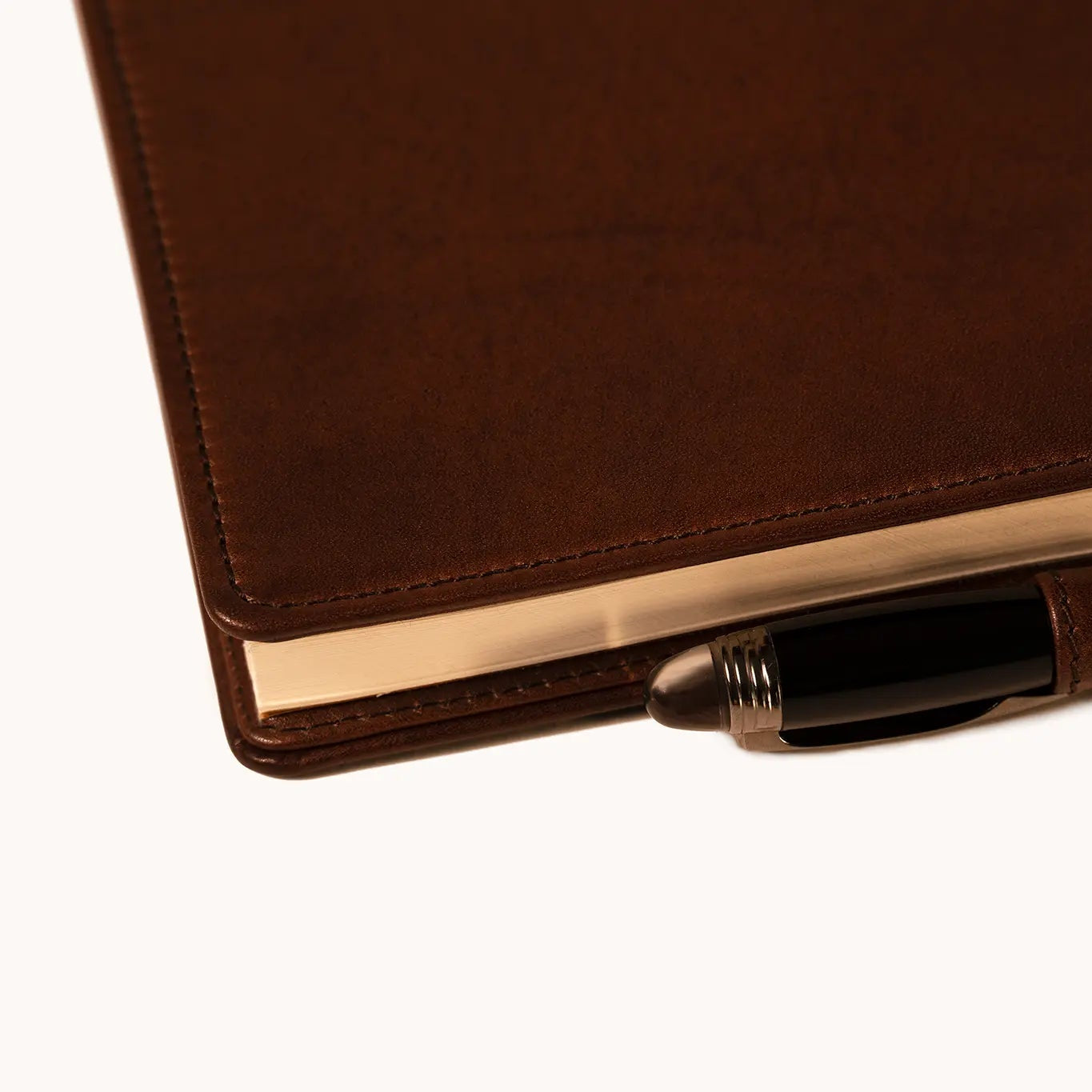 Lunburg Memoir Notebook Cover with Pen Loop in Deep Mahogany with an A5 notebook inserted and a pen held in the leather loop, resting on a table at a three-quarter angle showing the cover's profile, spine, and page block, in vegetable-tanned Perpetual Leather (from Tempesti) with folded edge construction.