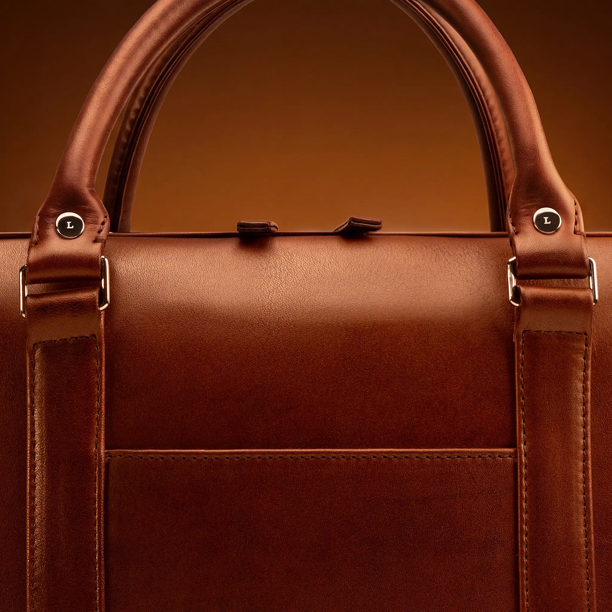 Close-up of the Lunburg Opus Briefcase handle and L-branded 316L stainless steel studs in Deep Mahogany Perpetual Leather (from Tempesti), the result of an object no machine and few masters can produce, showing folded edge construction and saddle stitching.