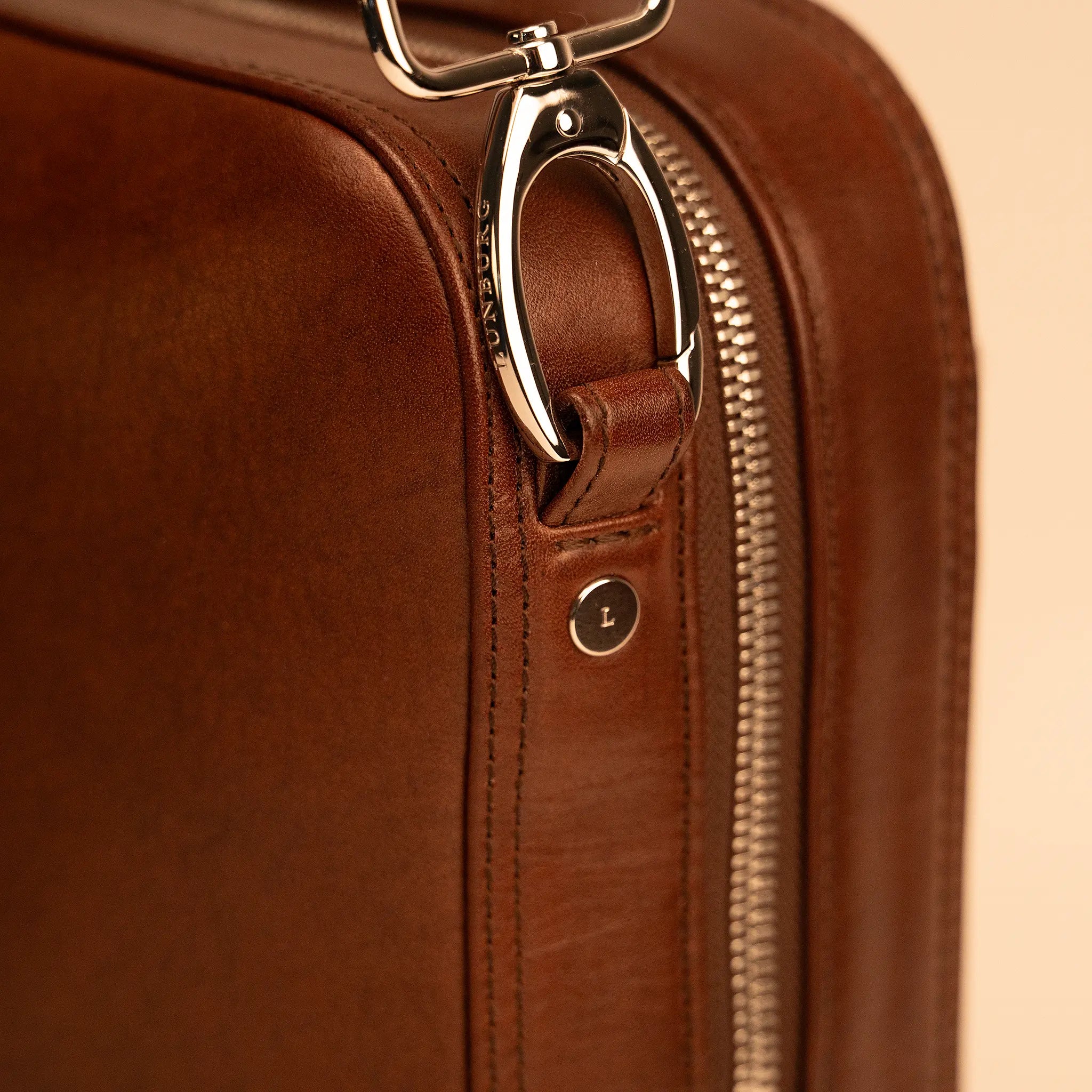Detail of the Lunburg Opus Briefcase showing the Lunburg-engraved shoulder strap clasp, L-branded stud, and YKK Excella zipper in 316L stainless steel, hardware engineered for silence against Deep Mahogany Perpetual Leather (from Tempesti) with folded edge construction.