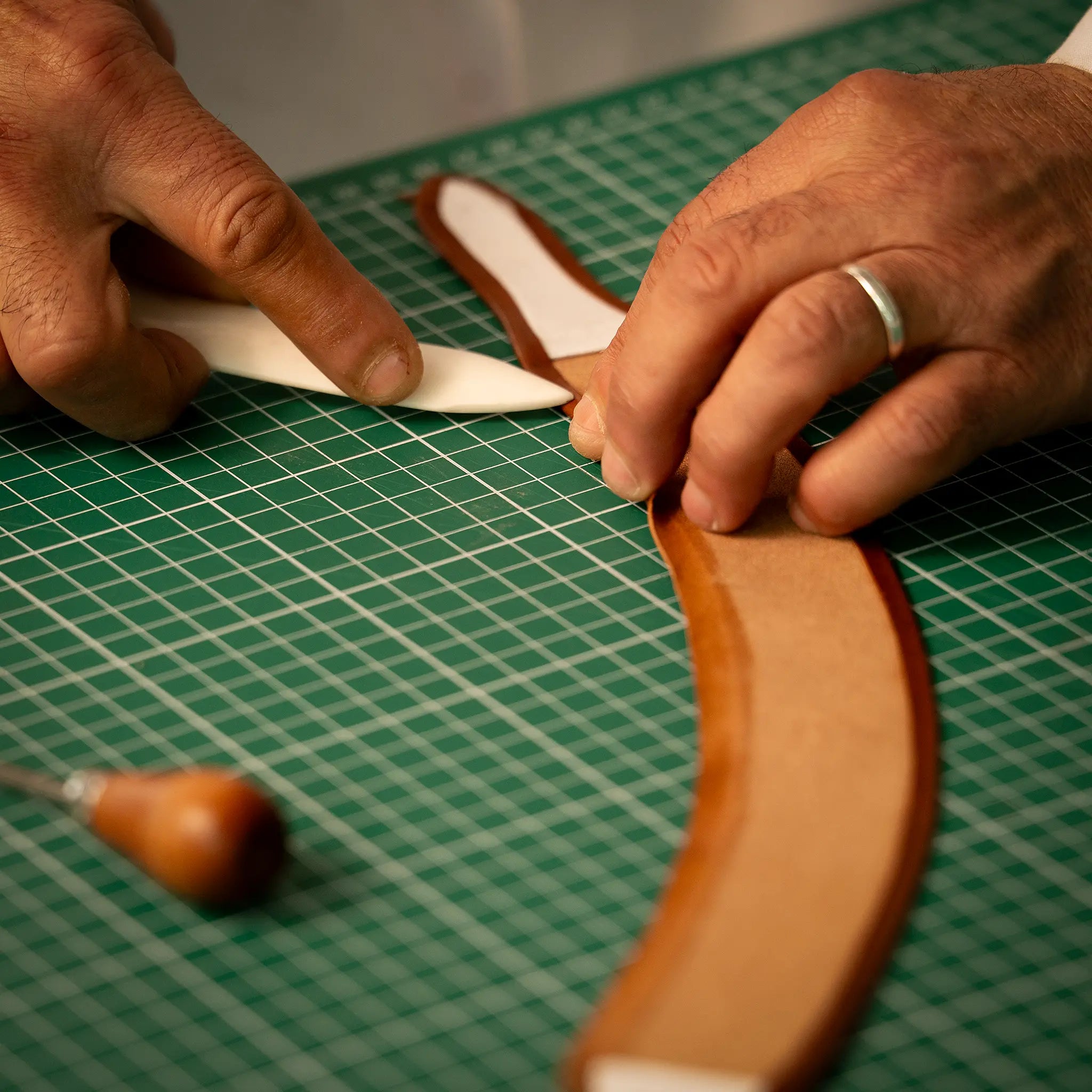 Close-up of artisan's hands using a bone folder to fold the leather edge during Rempliage at the Lunburg atelier, folding vegetable-tanned Perpetual Leather back upon itself on a cutting mat with an awl nearby.