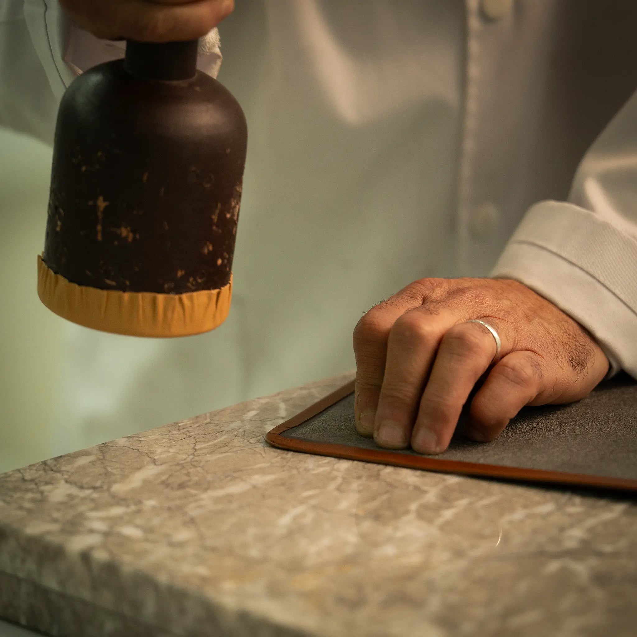 Artisan's hand holding the rempliage hammer poised above a folded edge leather panel on the marble slab at the Lunburg atelier, setting the dead edge where strike pressure and angle are calibrated to the temper of the hide.