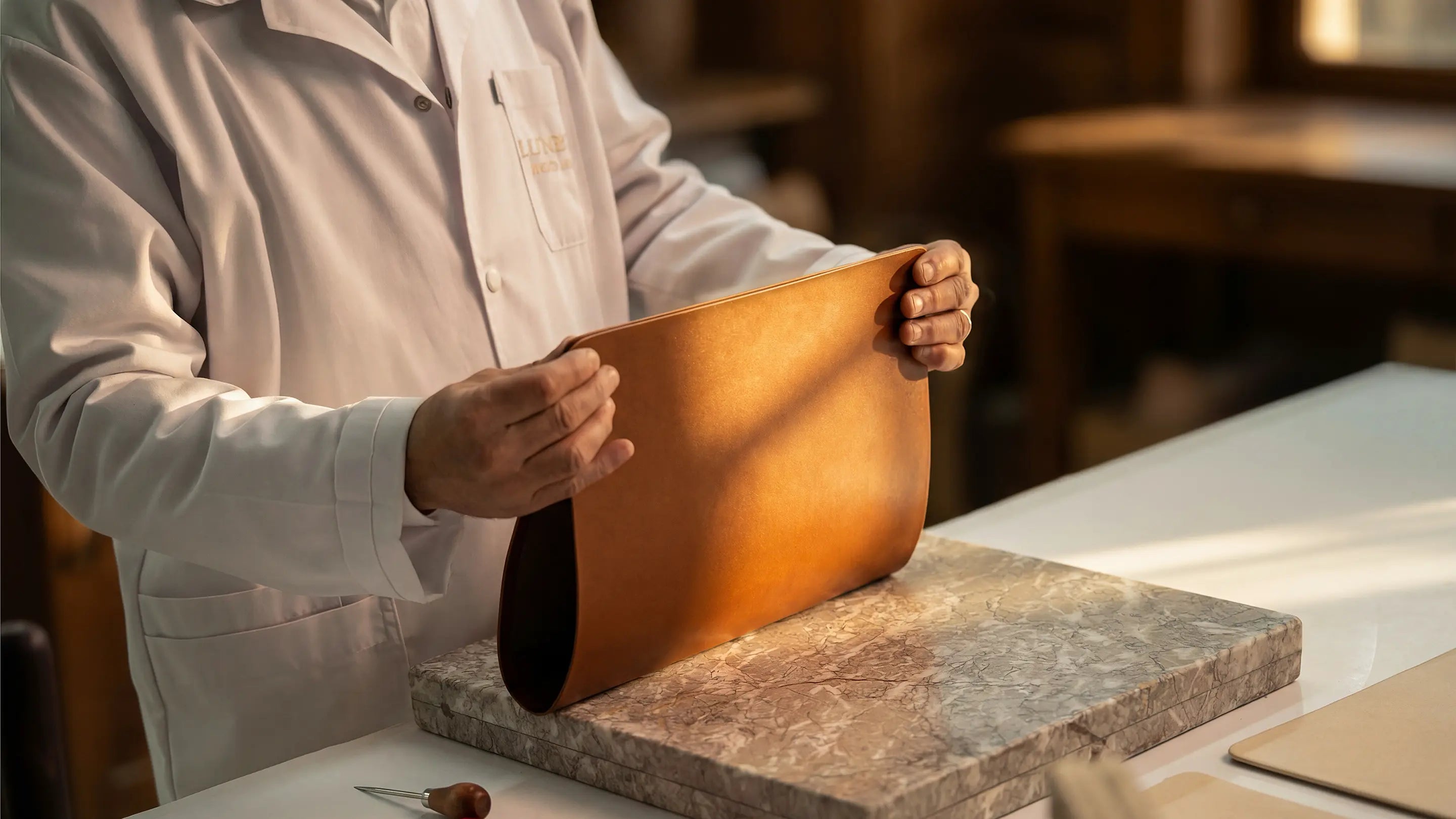 Artisan at the Lunburg atelier holding a vegetable-tanned Perpetual Leather panel above a marble slab, inspecting the folded edge of Rempliage where the grain curves seamlessly from exterior to interior with no visible seam.