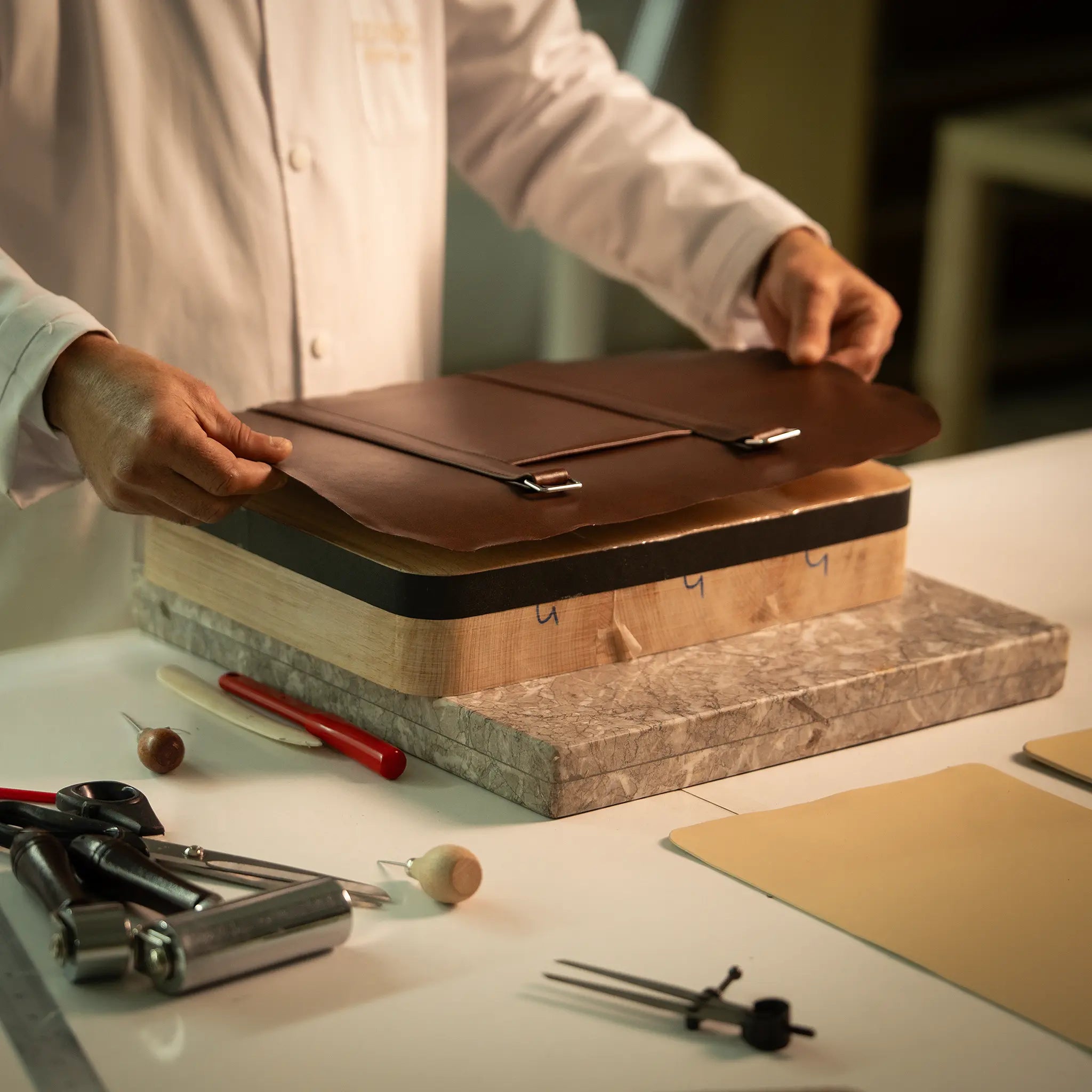 Artisan placing a leather panel onto the forme en bois wooden last at the Lunburg atelier, with traditional tools including bone folder and marble slab visible, assembling layers for the Rempliage folded edge construction.