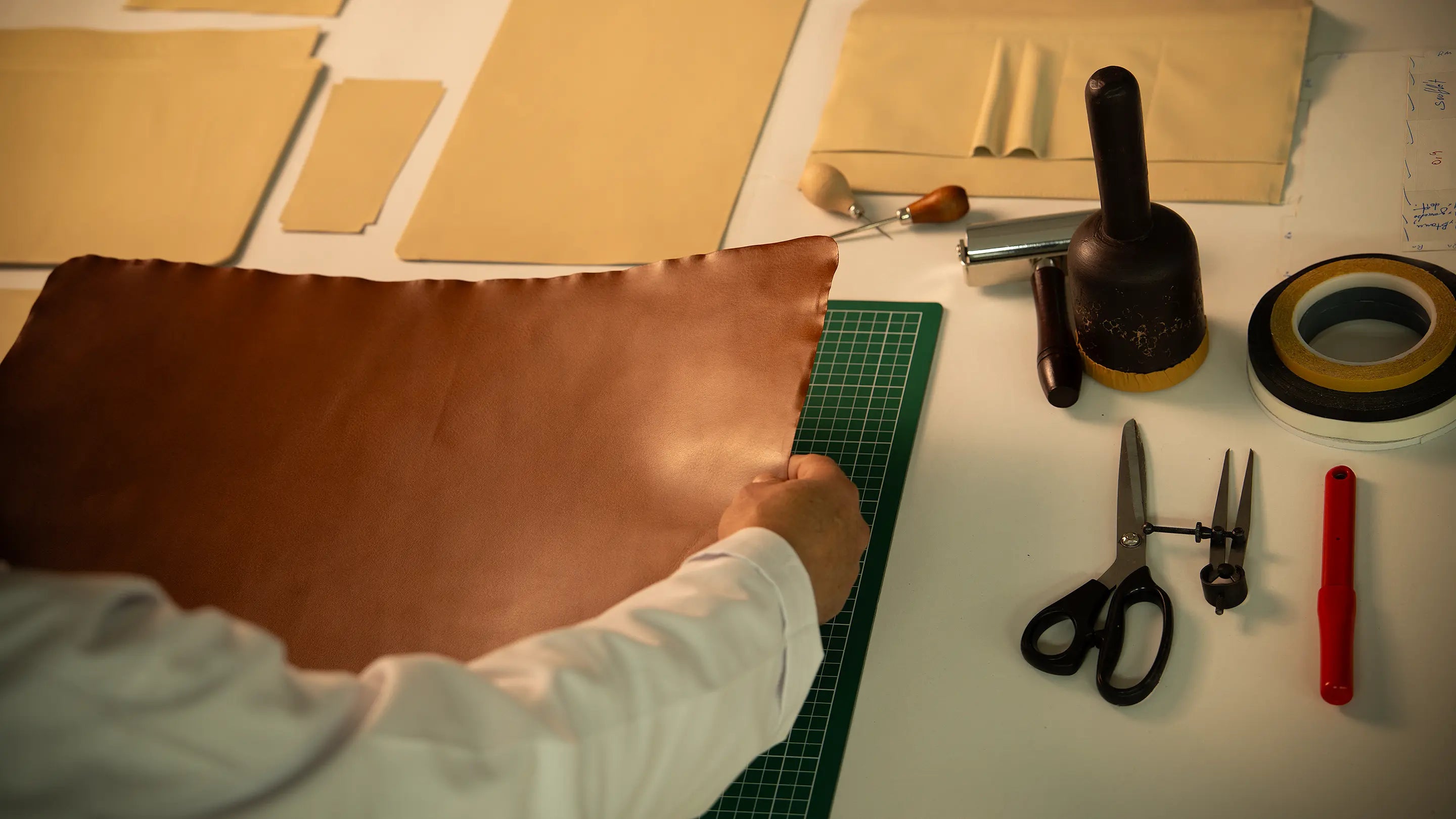 Overhead view of the Lunburg atelier workbench during Rempliage preparation, with an artisan's hand holding a vegetable-tanned Perpetual Leather panel alongside working mat, pattern templates, rempliage hammer, and traditional leatherworking tools.