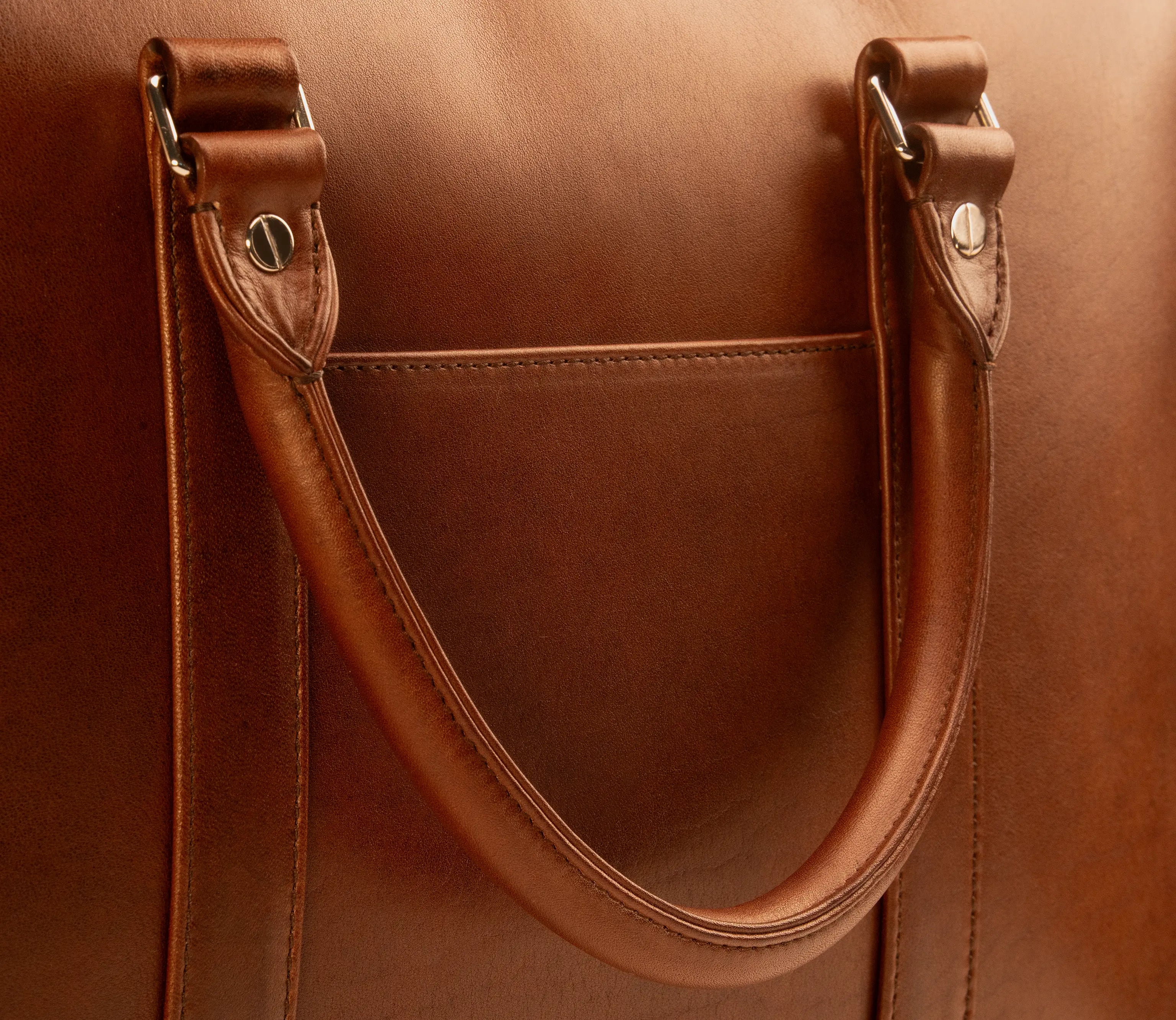 Close-up of the Lunburg Opus Briefcase handle attachments and rear slip pocket, showing the seamless folded edge of Rempliage where leather grain flows continuously from exterior to interior, in vegetable-tanned Perpetual Leather with 316L stainless steel hardware.