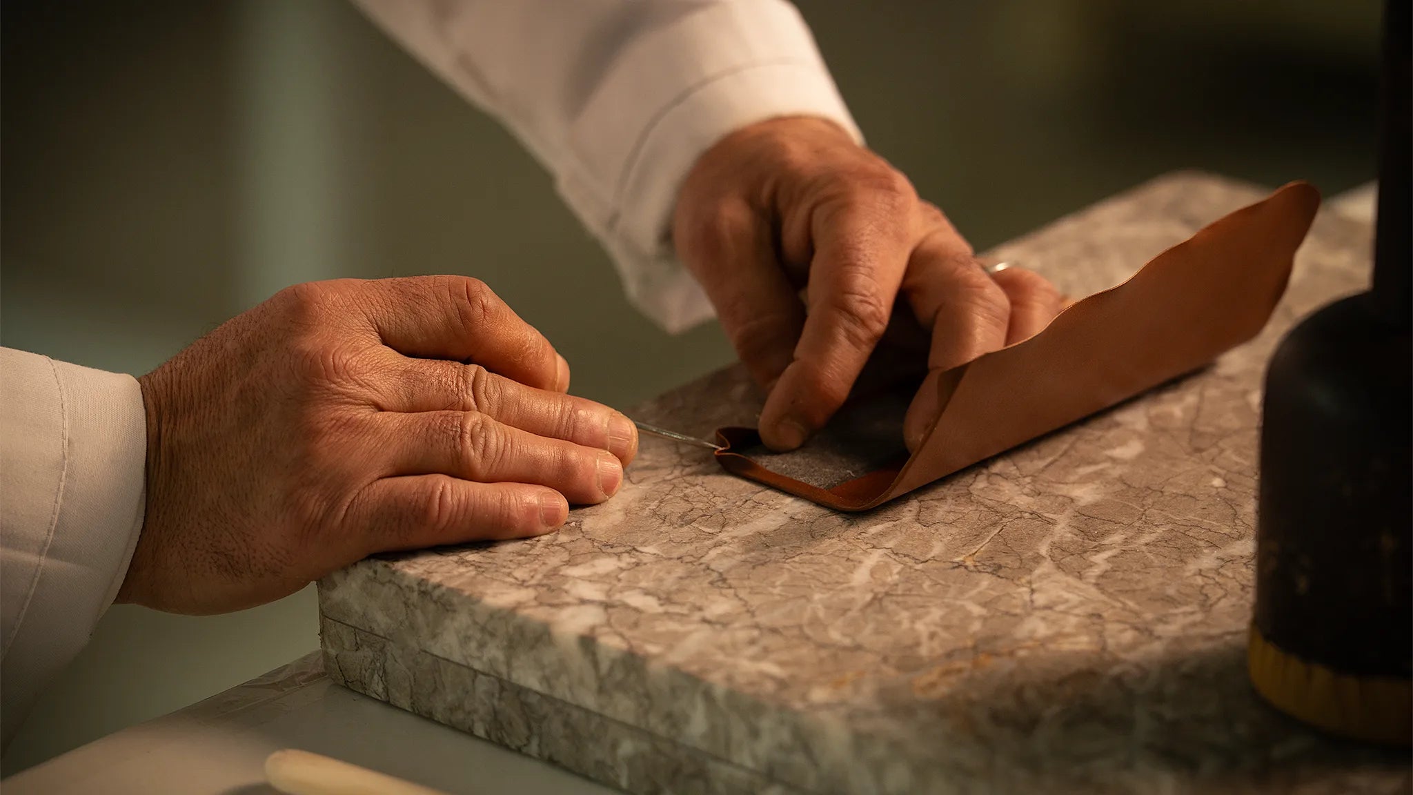 A Master Artisan's hands at the Fes atelier performing Rempliage on a small panel of vegetable-tanned Perpetual Leather (from Tempesti), folding the edge back upon itself against a marble work surface, demonstrating the discipline of restraint where every fraction of a millimetre is final and the smaller the object, the more unforgiving the execution.