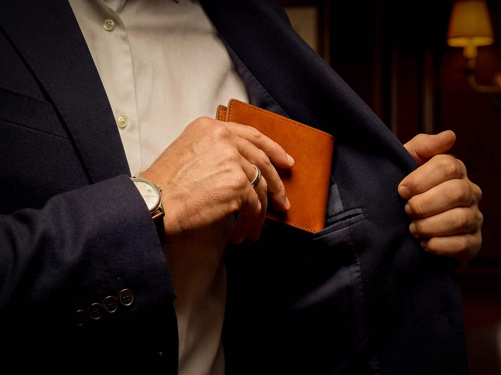 A hand slipping a Lunburg Signet wallet in Heritage Amber vegetable-tanned Perpetual Leather (from Tempesti) into the interior breast pocket of a navy suit jacket, demonstrating the wallet's slim profile and folded edge construction refined enough to disappear beneath fabric, craft that serves without announcing itself.