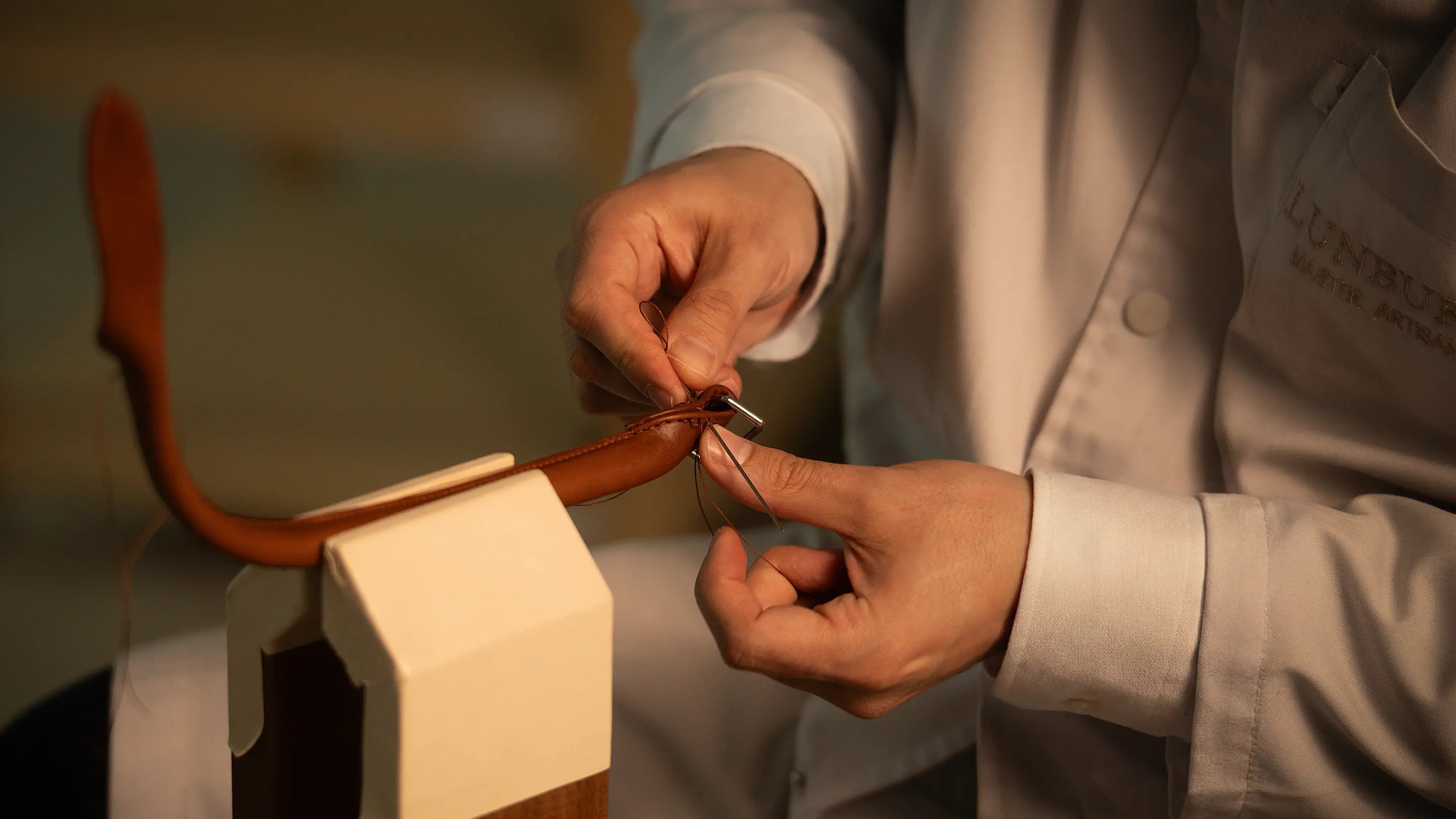 Artisan's hands at the Lunburg atelier hand-stitching a leather handle on a stitching pony, with two needles drawing thread through vegetable-tanned Perpetual Leather, the structural craftsmanship behind the Lunburg Fifty-Year Warranty.